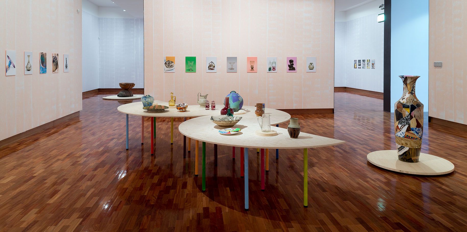 Plates bowls and decorative objects made from various materials are displayed on a plywood table with colourful prints hanging on the wall behind. A large human height colourful vase is positioned next to the tables on a low plinth,