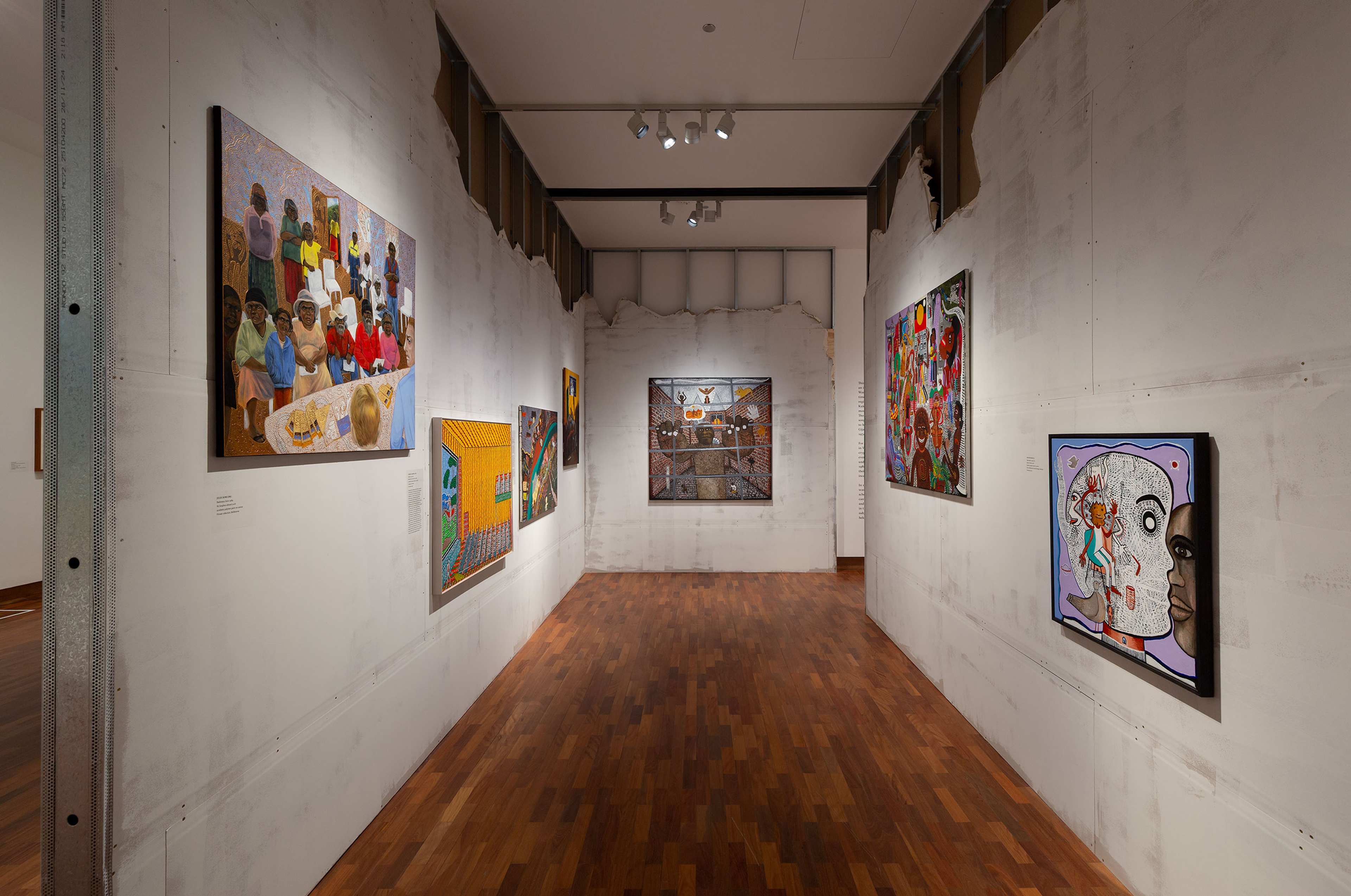 Art gallery with wooden floors and white walls displays colourful paintings, including portraits and abstract art, hung on both sides and at the end of a well-lit, narrow hallway.