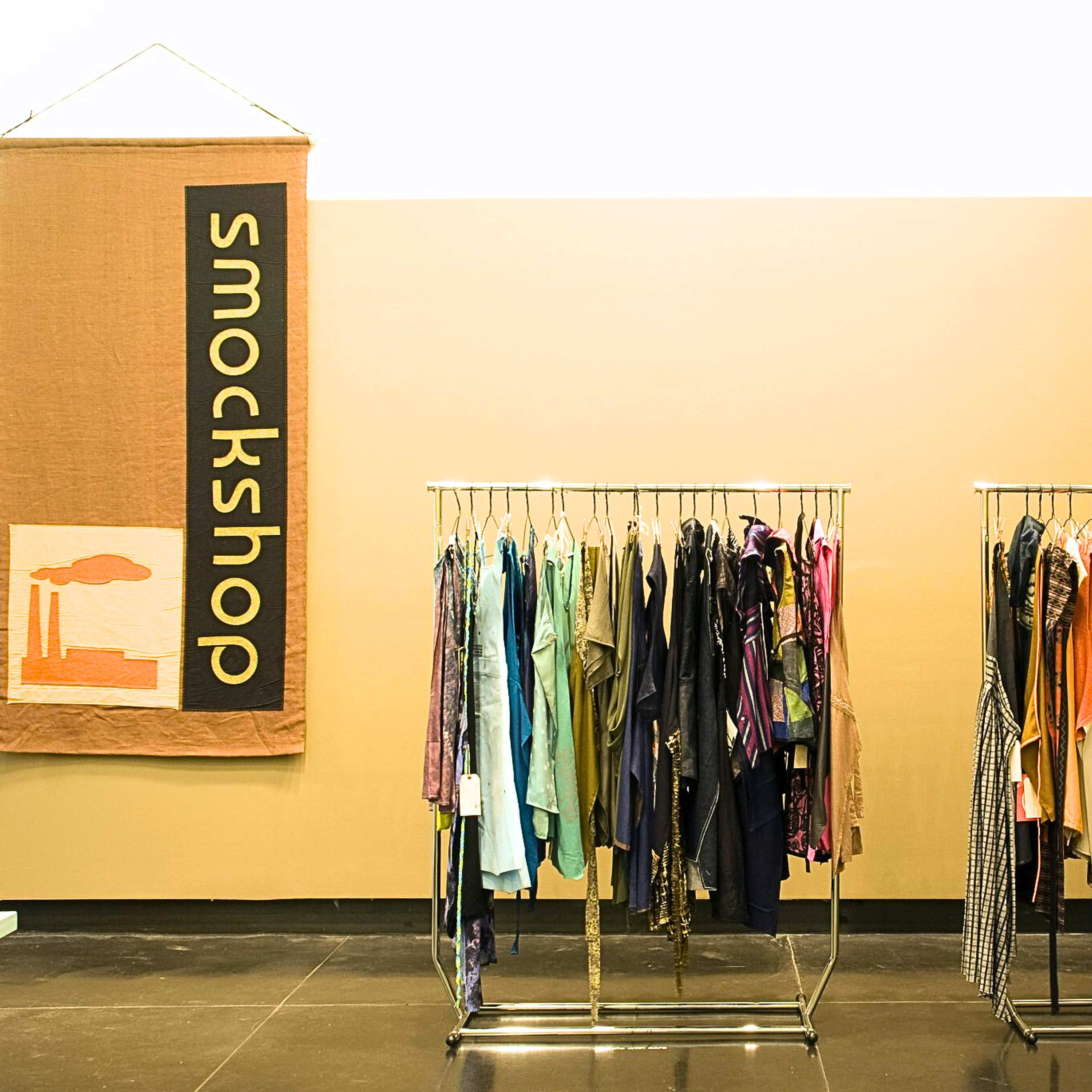 A photograph of a gallery with two clothing racks full of dresses and a large sign that says 'smockshop' on the left