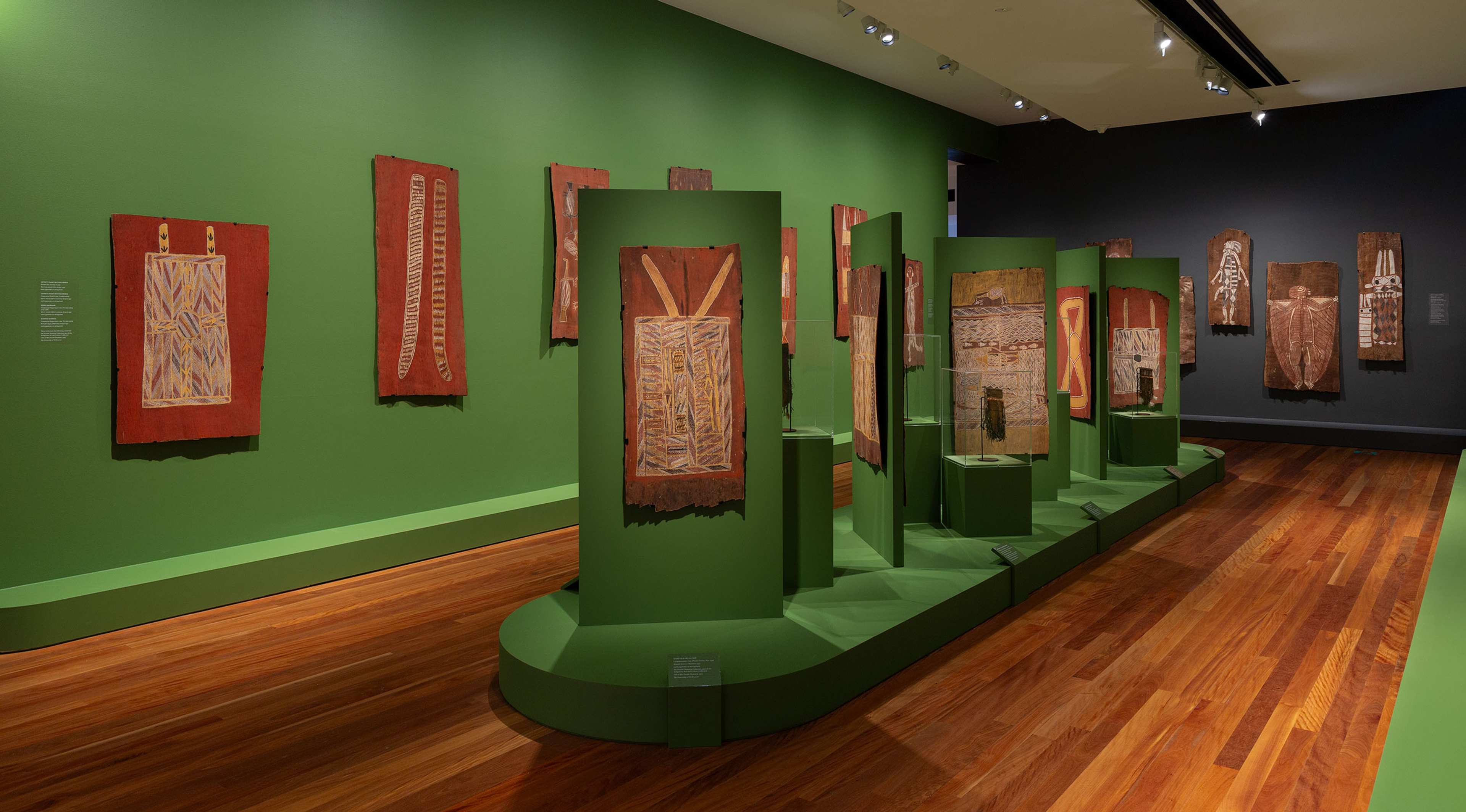 A gallery with green walls and wooden floors displays bark paintings, some hung on the walls and others on green pedestals in the center. The artworks feature traditional Indigenous patterns and earthy colors.