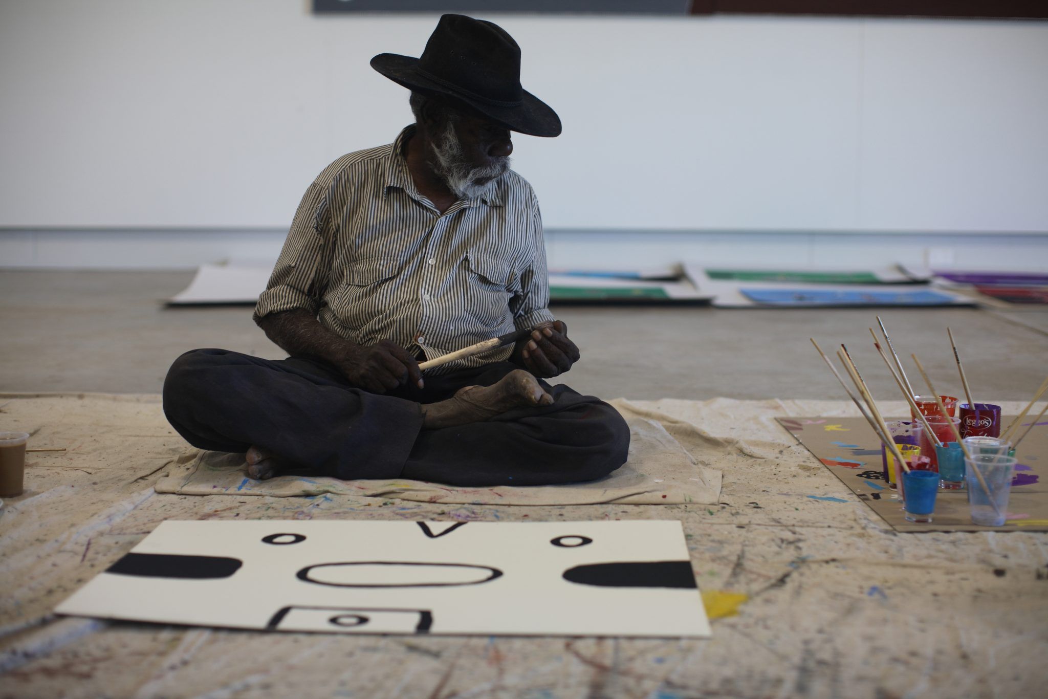 A photograph of a black person in a wide-grinned hat sitting cross-legged on the floor of a room with a black-and-white geometric painting in front of them and some jars holding brushes to their left 