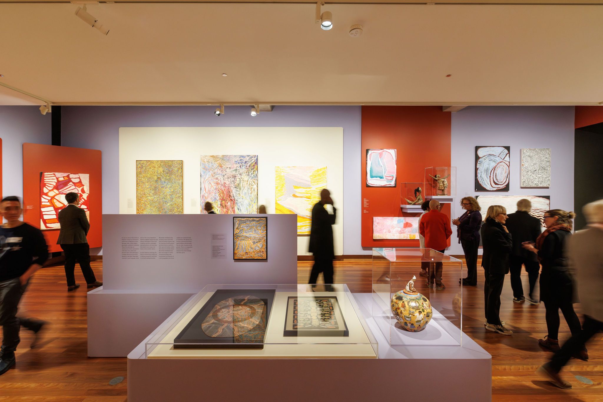 A brightly lit art gallery with people viewing paintings and sculptures. Colorful abstract artworks hang on red and blue walls, and glass display cases hold various items on wooden floors.