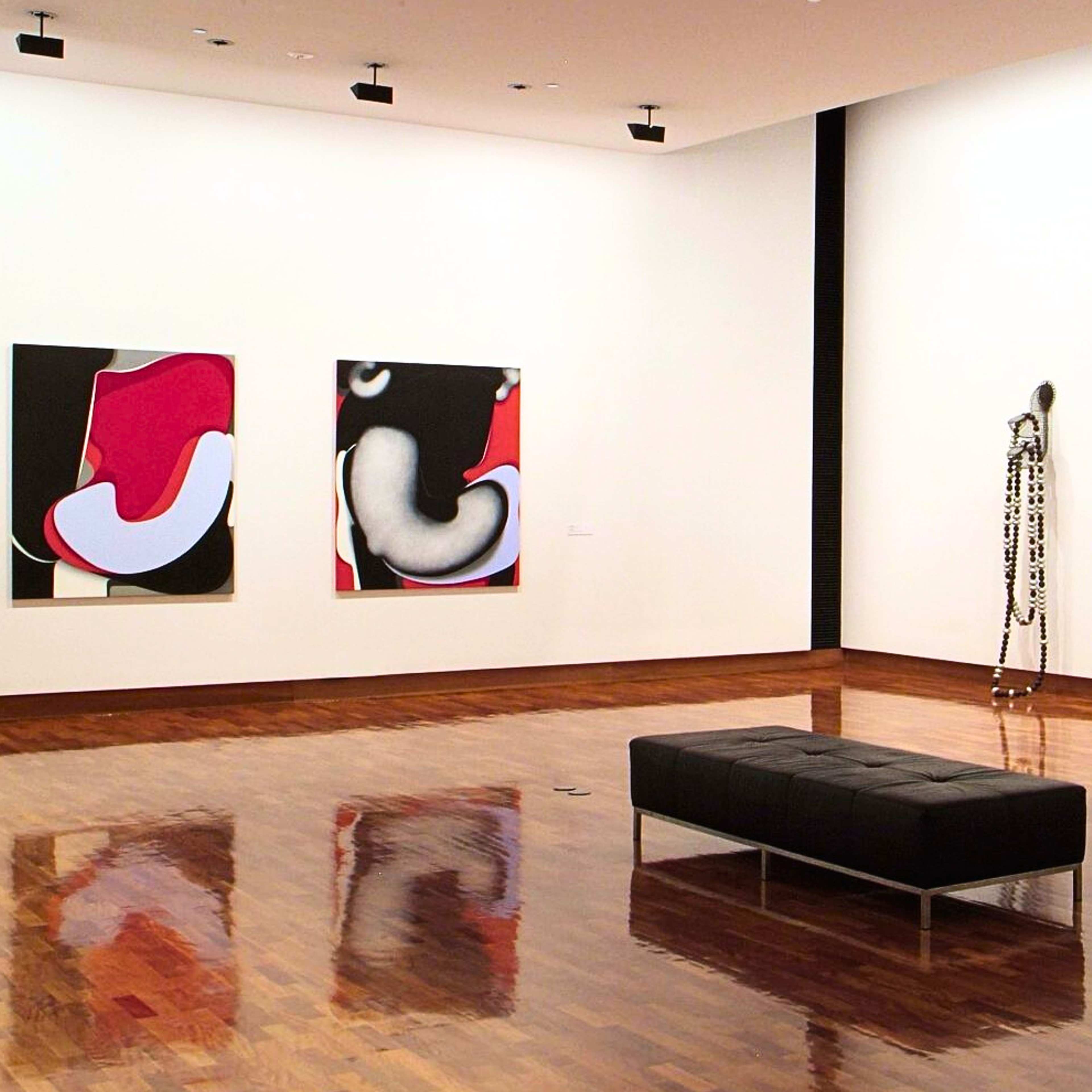 A photograph of a gallery space with a row of four sculptures reminiscent of giant necklaces on one wall and two square red and black images on the other