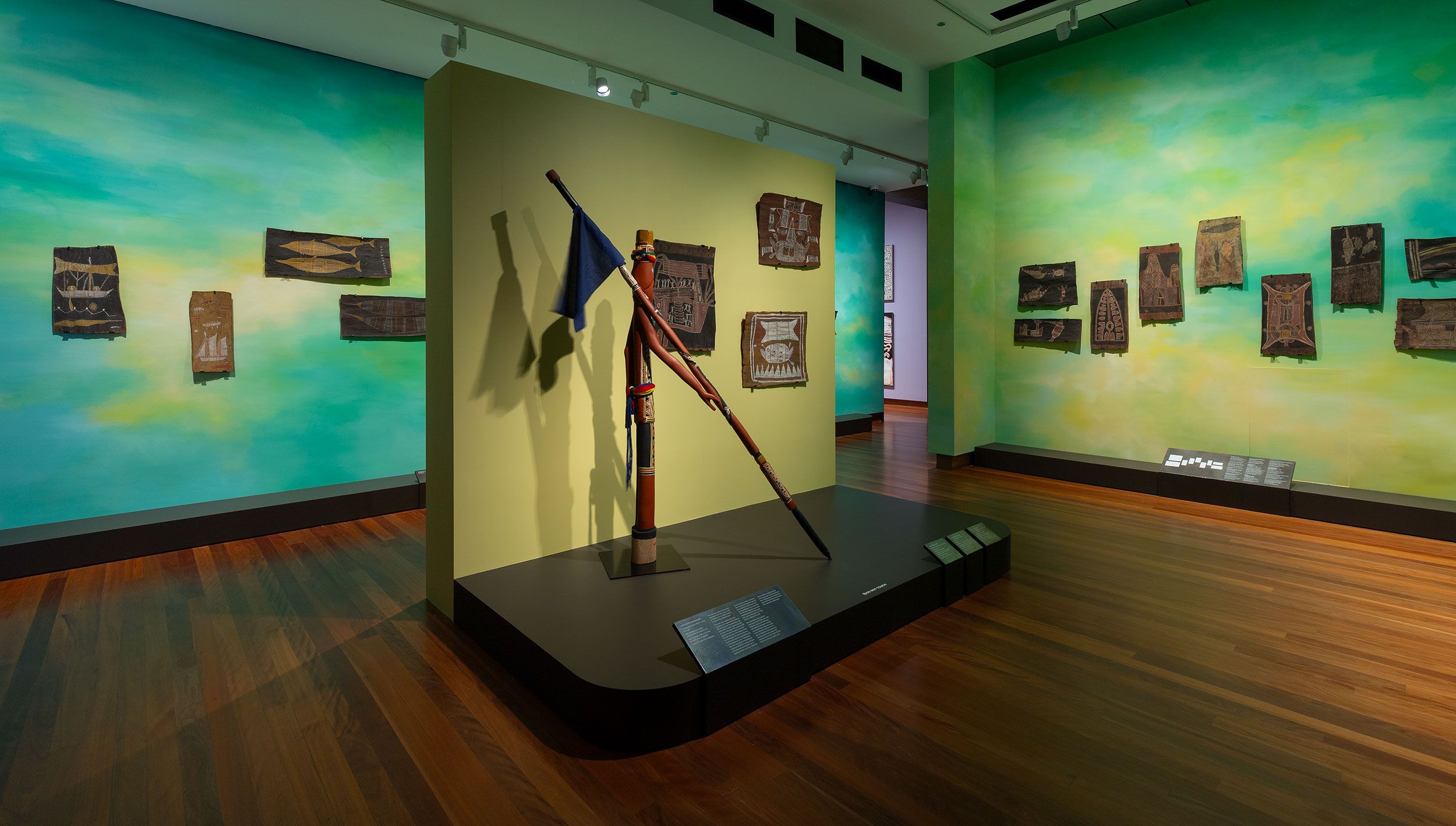 A museum exhibit features wooden artifacts mounted on colourful turquoise and green walls, with two large carved wooden spears displayed upright on a central platform on polished wooden floors.