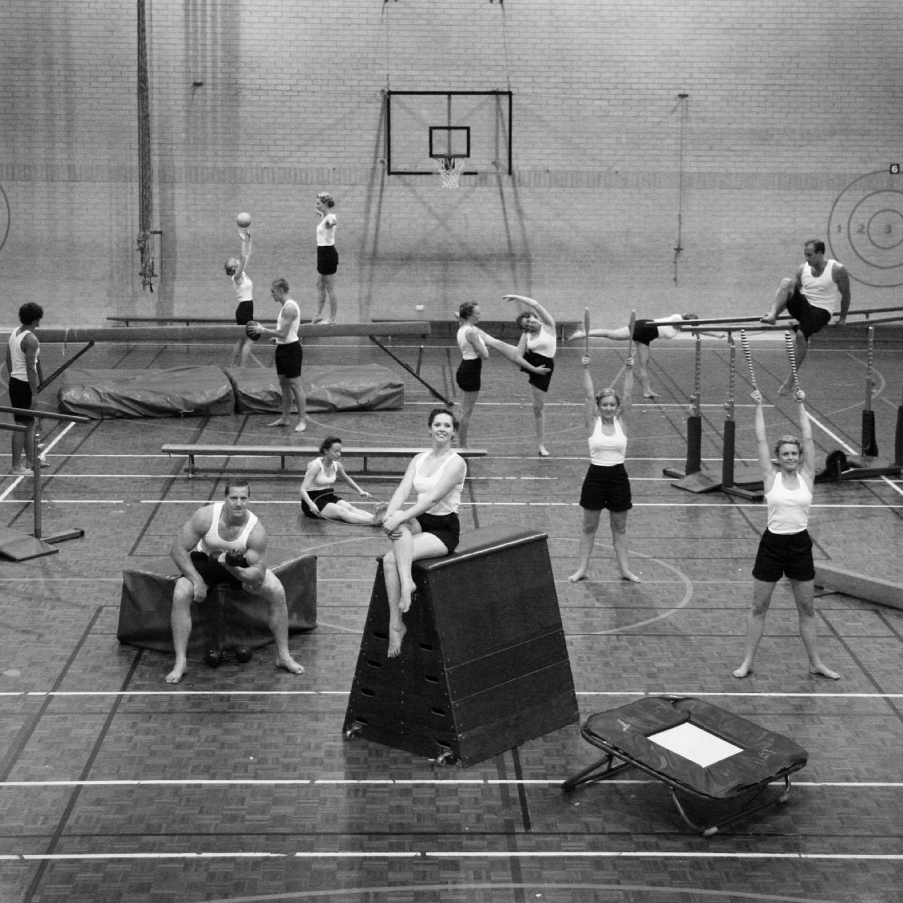 A black and white photograph of people wearing black shorts and white singlets exercising in an old-fashioned gym