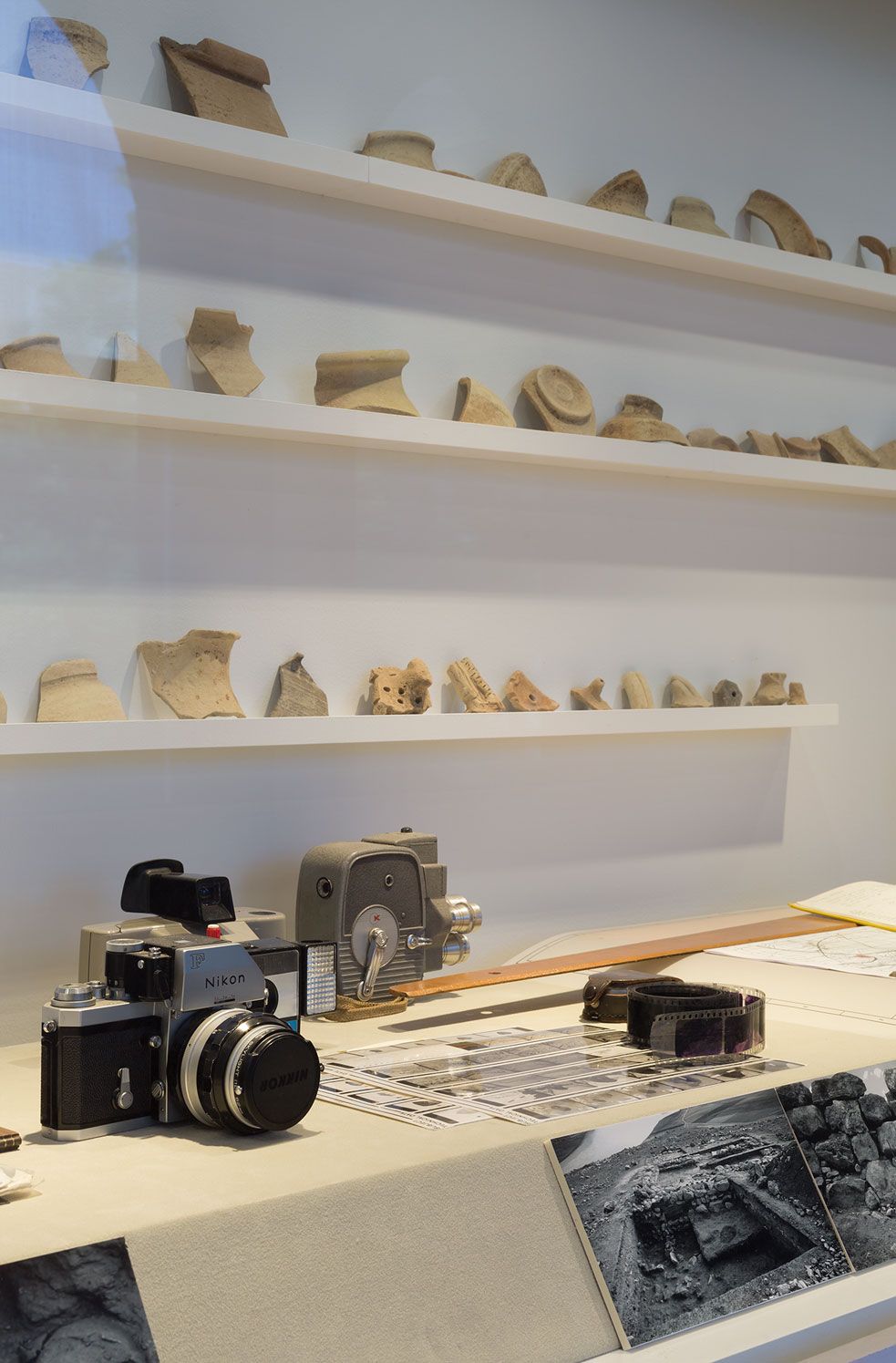 A photograph of a gallery with a large vitrine displaying a large array of ancient ceramics and a film photographic camera and some black and white photographs in the foreground