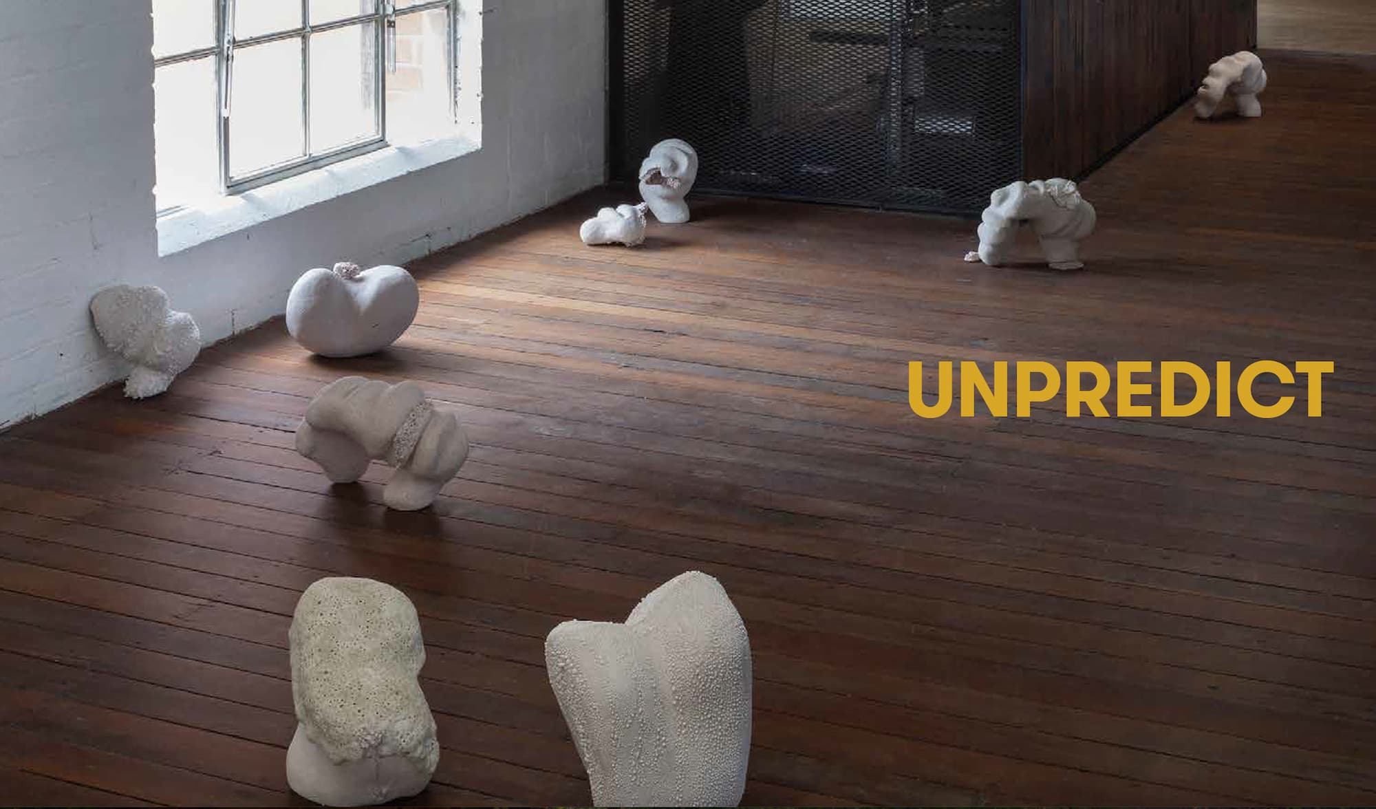 White abstract sculptures are scattered across a wooden floor near a large window, with the word “UNPREDICT” in bold yellow text on the right side of the image.