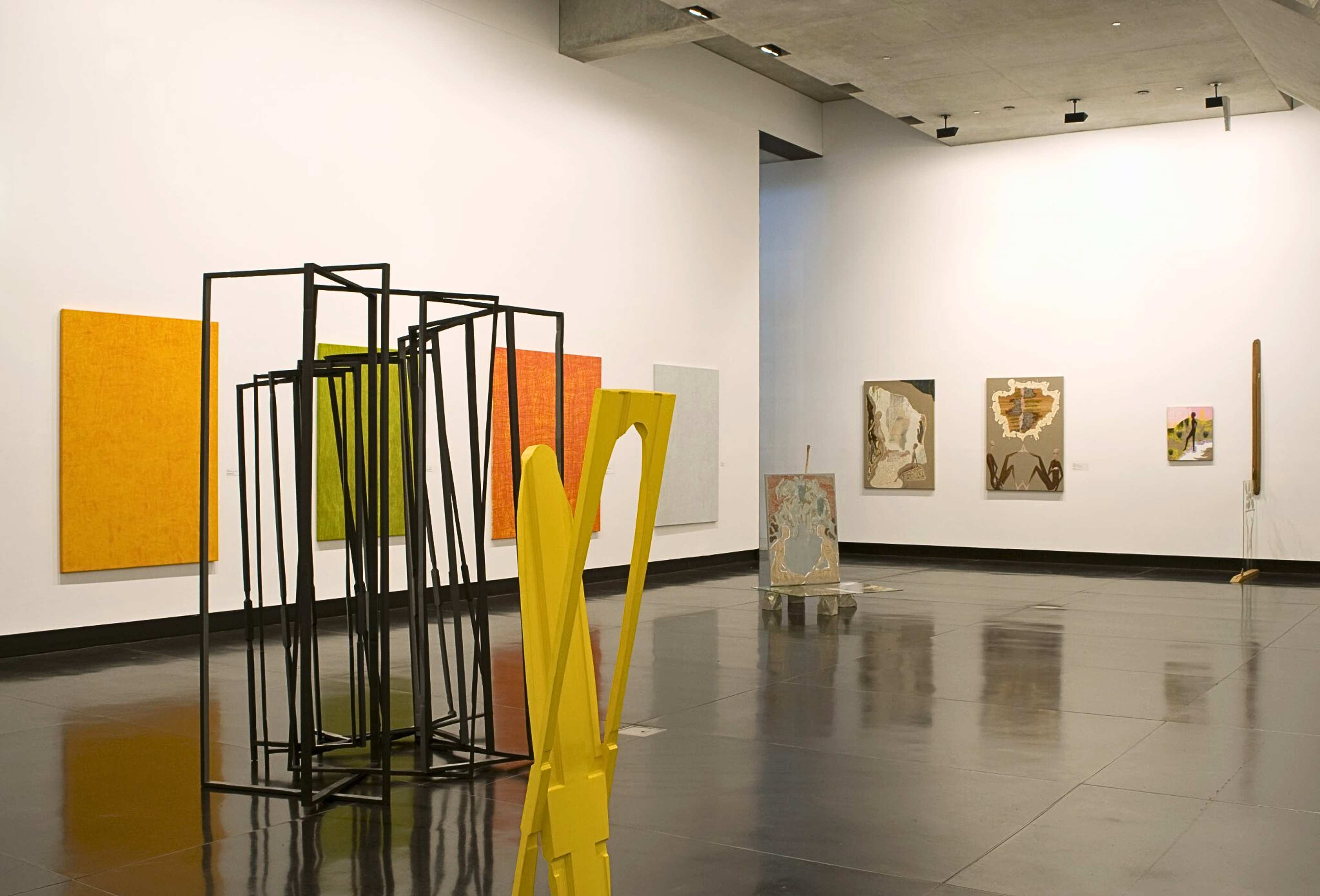 A photograph of a gallery with large sculpture consisting of several frames in the foreground and several colourful paintings behind it