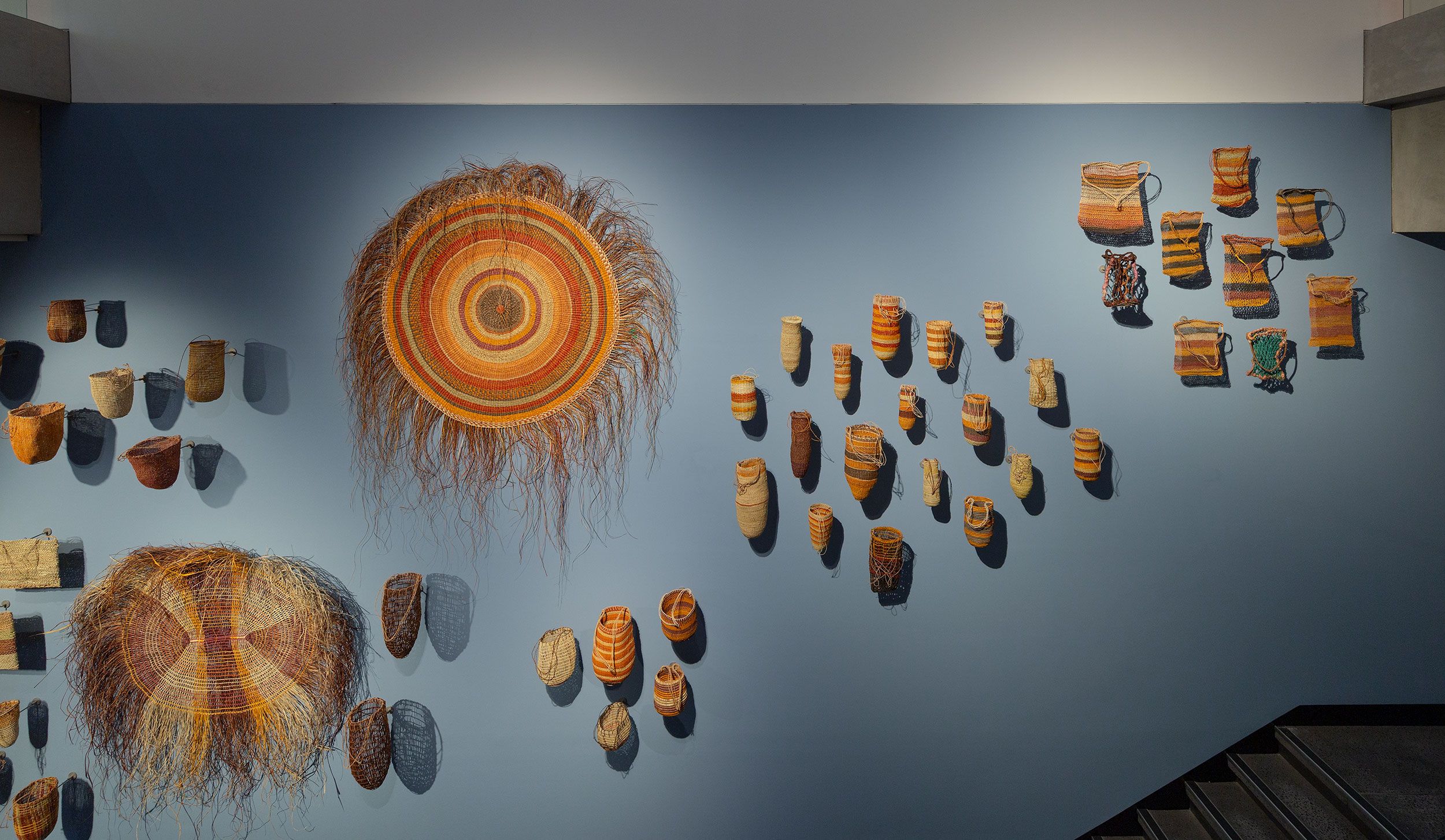 Woven baskets and large, circular woven art pieces are displayed on a blue wall, arranged in clusters and rows, with stairs visible at the bottom right corner.