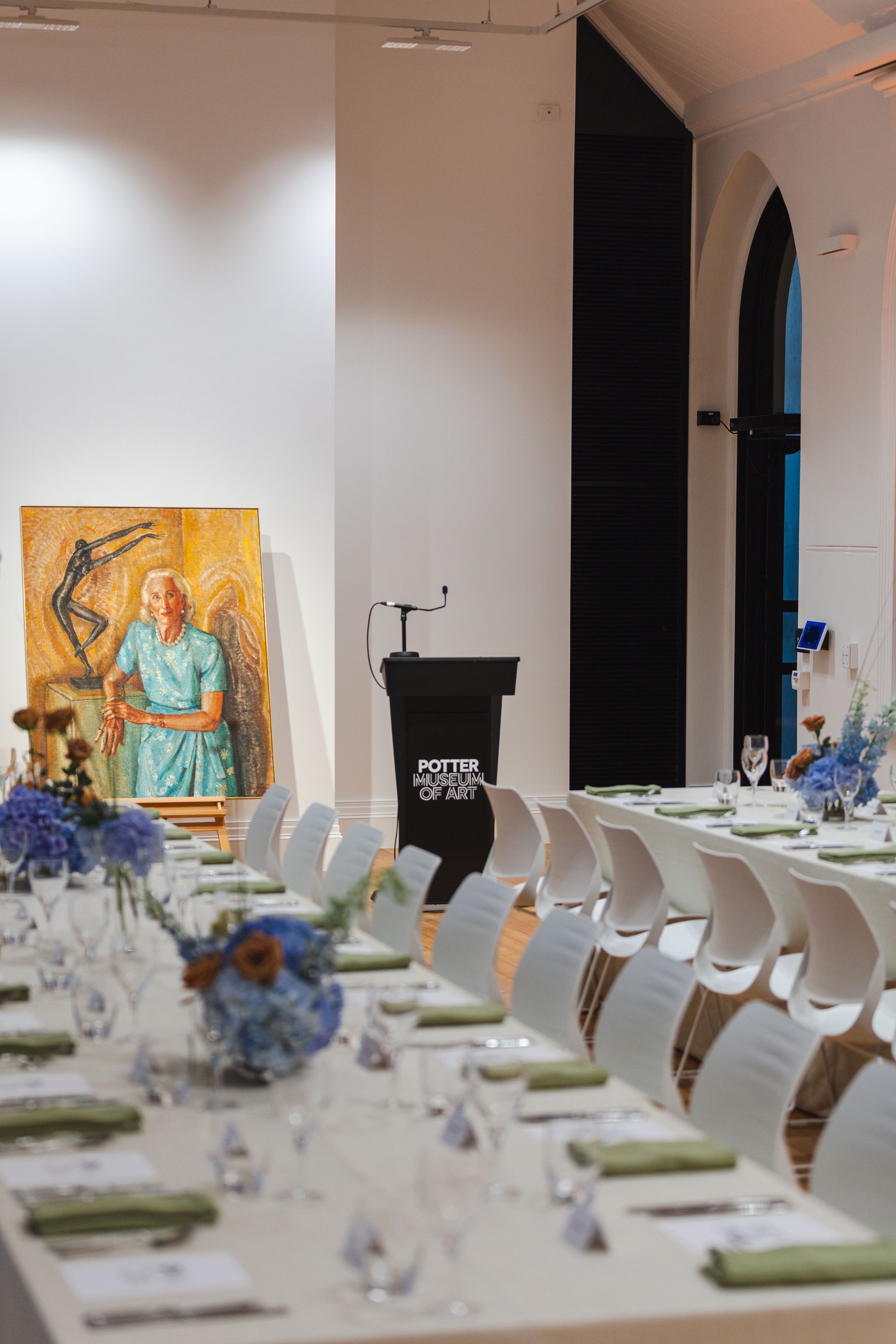A formal dining setup with white chairs and tables decorated with blue flowers, positioned in front of a podium labeled Porter School of Art and a colourful painting of an elderly woman and a dancer.
