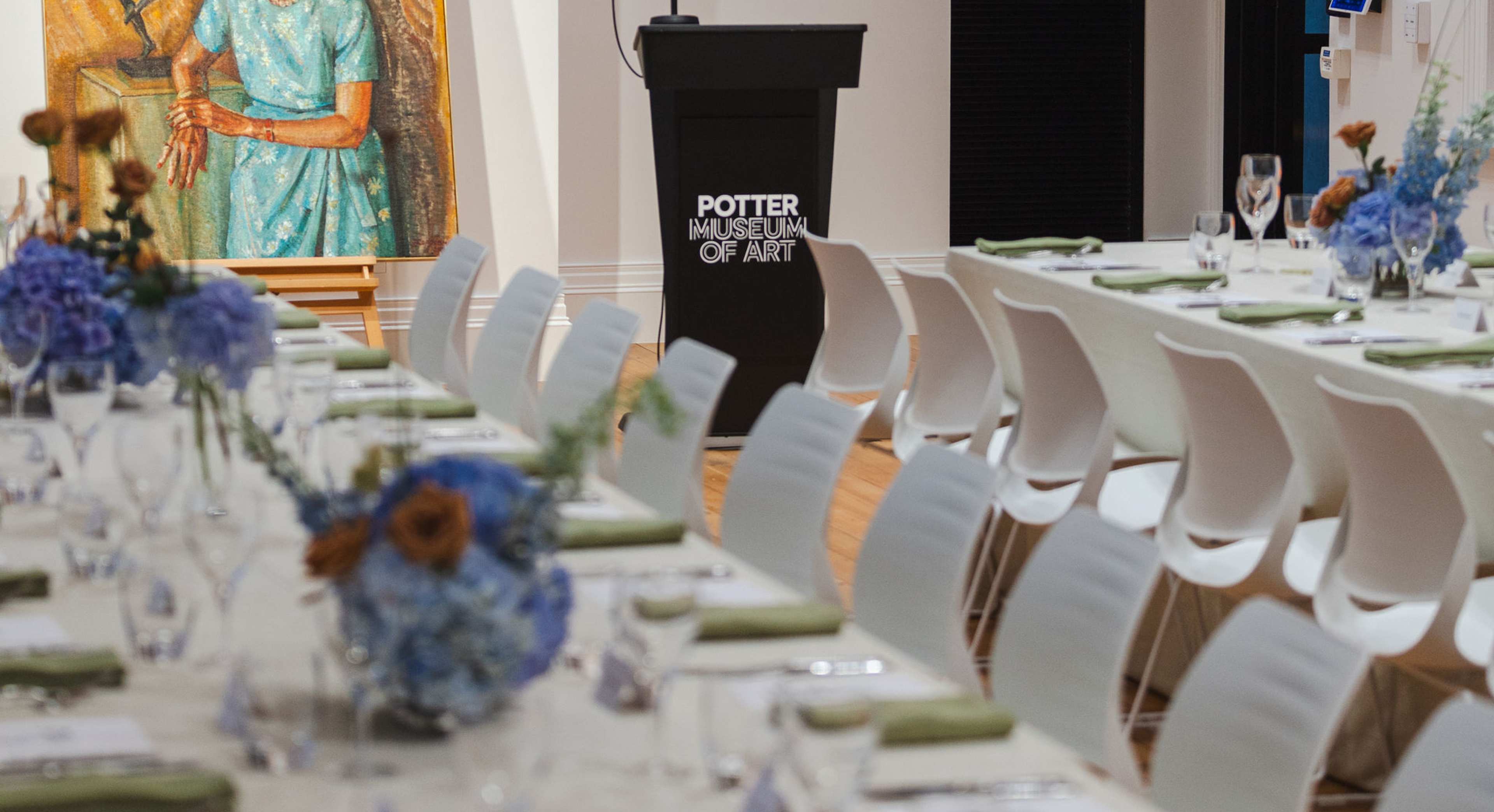 A formal dining setup with white chairs and tables decorated with blue flowers, positioned in front of a podium labeled Porter School of Art and a colourful painting of an elderly woman and a dancer.