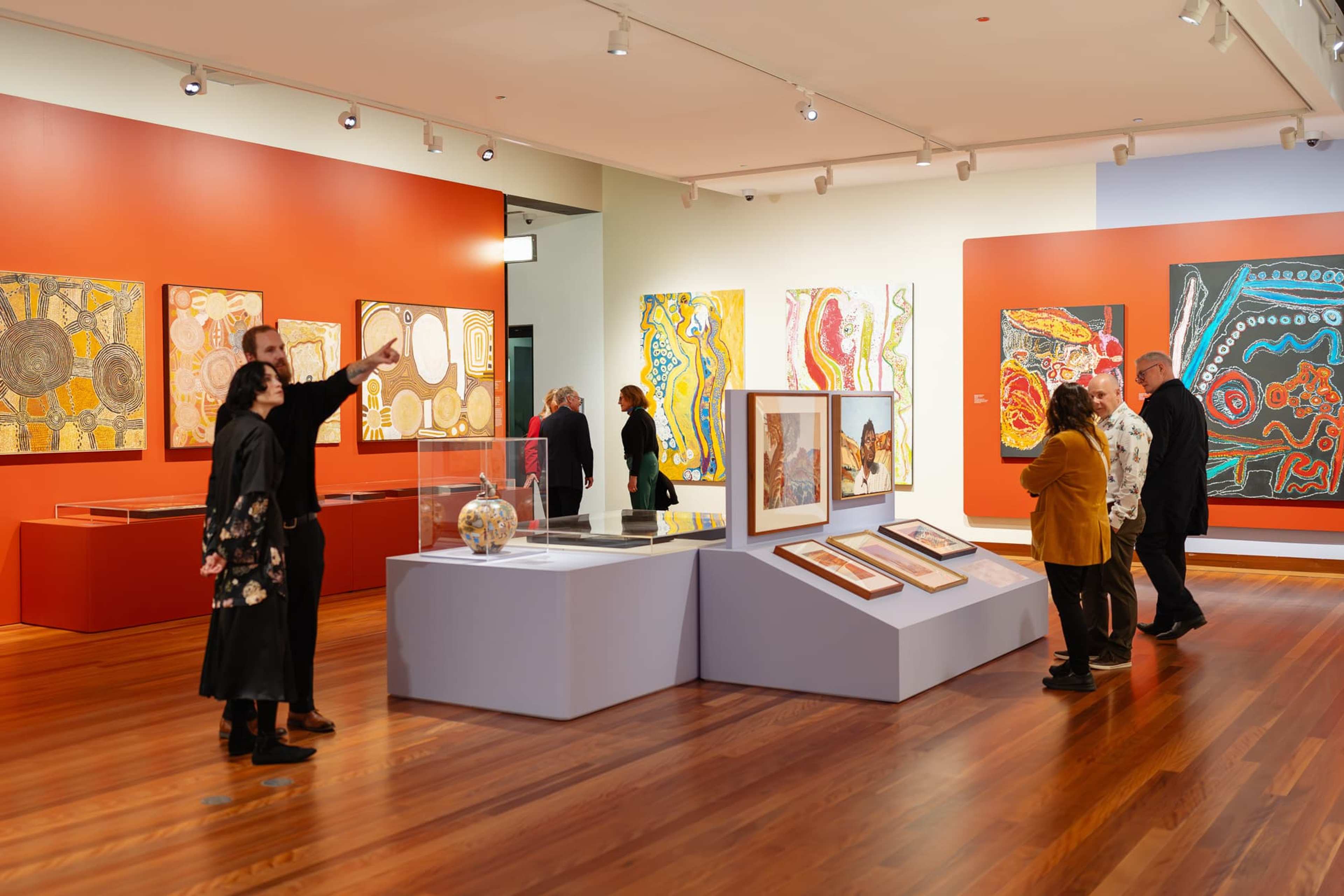 Visitors view and discuss colourful abstract artworks and artefacts displayed in a modern art gallery with red accent walls and wood floors. Some people read information panels, while others observe the exhibits together.