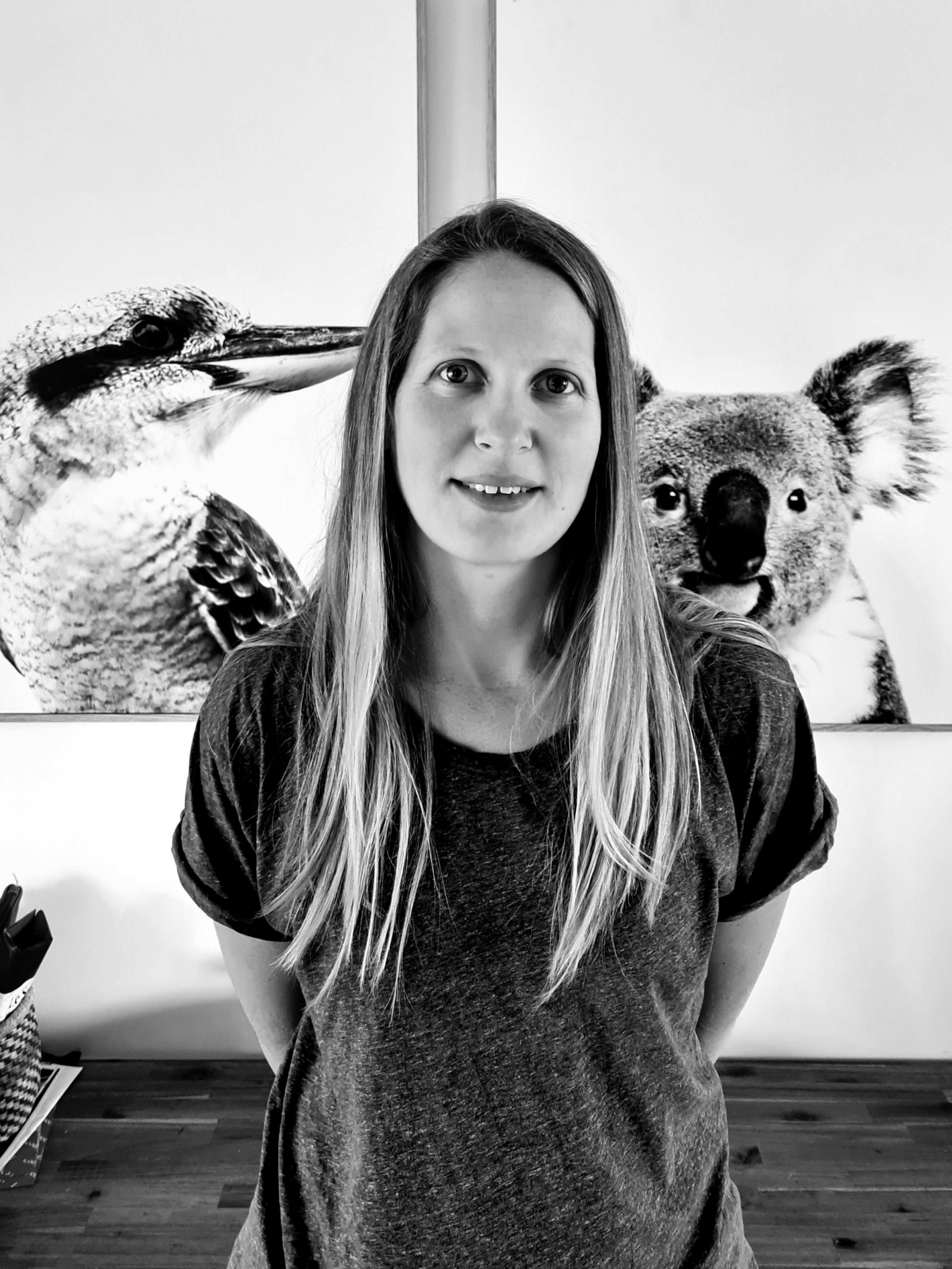 A woman with long hair stands indoors in front of two animal portraits, one of a bird and one of a koala. The image is black and white.