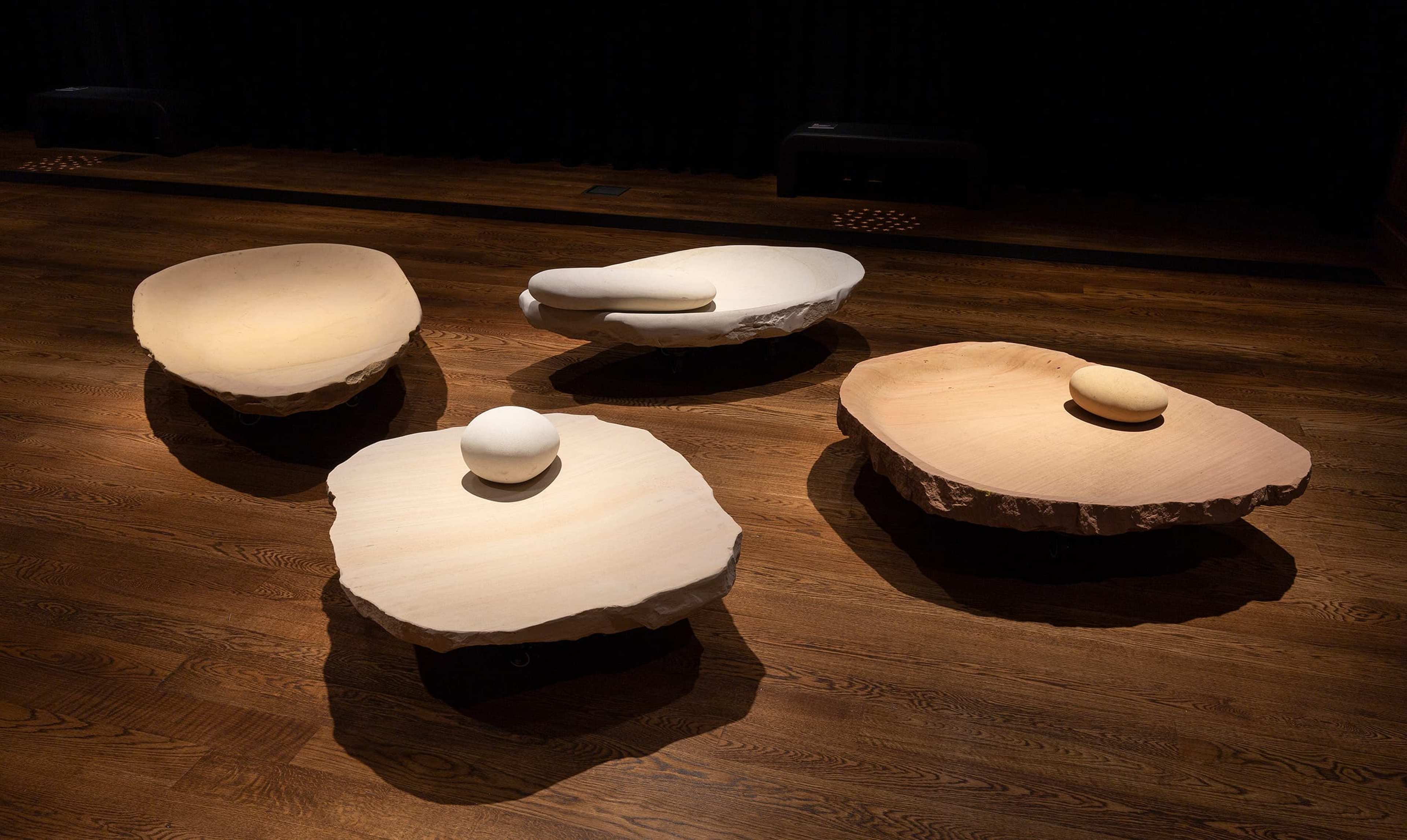 Four sculptural seats resembling smooth, organic stone shapes are arranged on a wooden floor; each piece has a unique form and natural, earthy color tones, with rounded cushion-like elements on top.