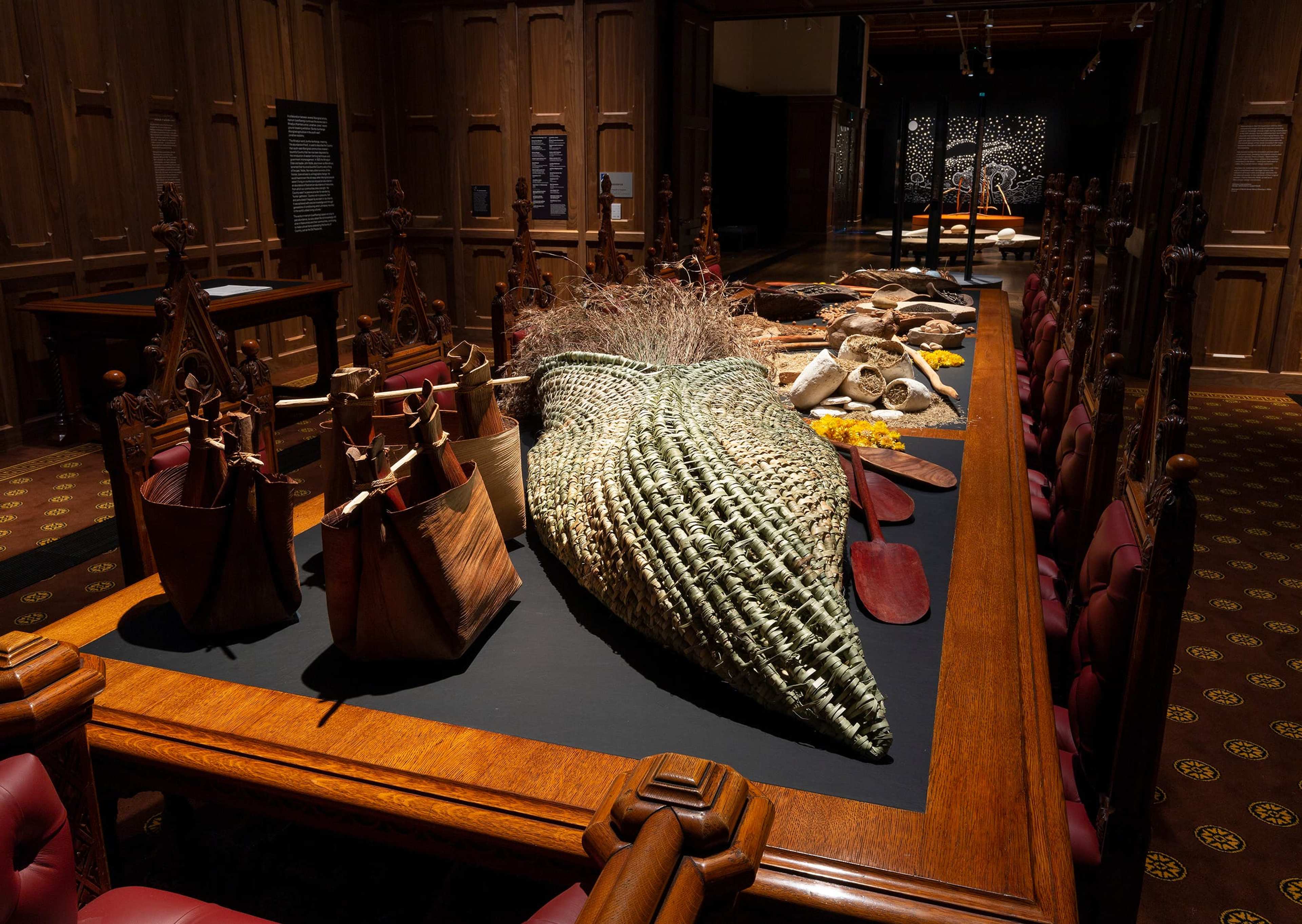 A long wooden table in a dimly lit room displays woven baskets, paddles, wooden bags, and other handcrafted items. The room has ornate wood paneling and carved chairs around the table.