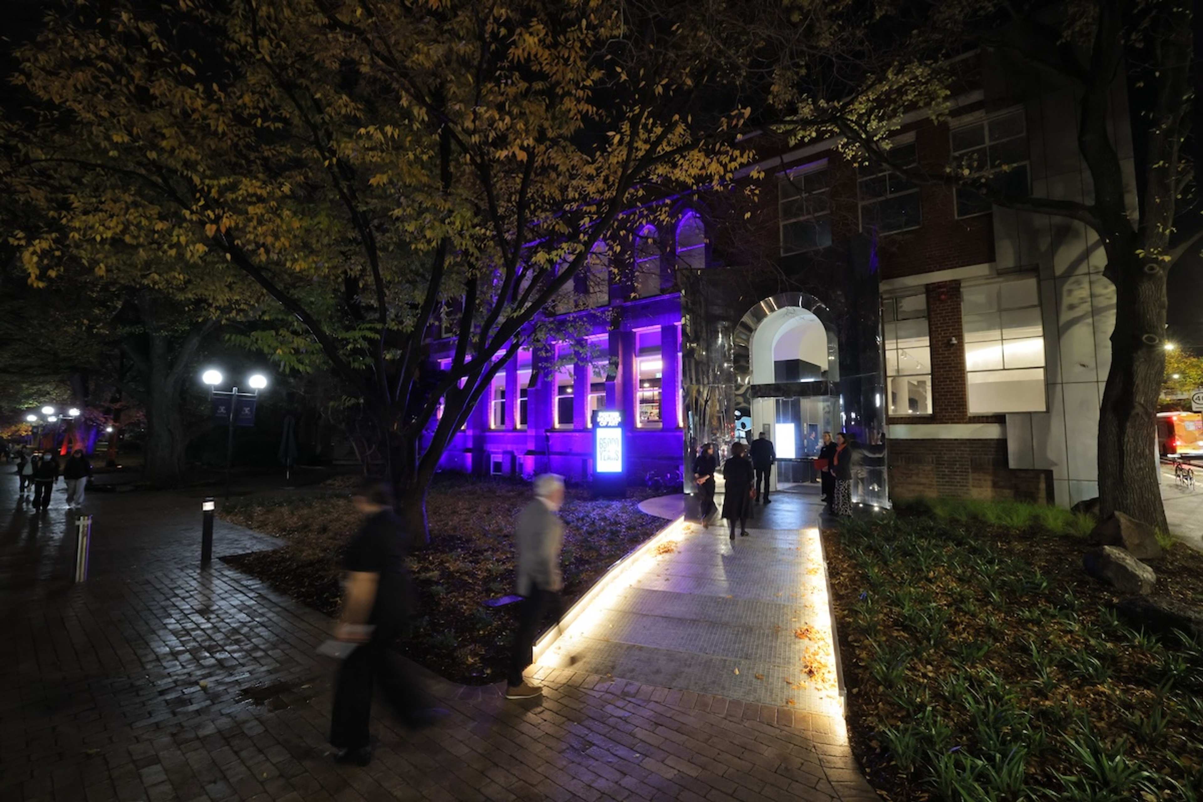 A group of people gather at an exhibition or event in front of a large wall display titled 65,000 Years: A Short History of Australian Art with text and flowers on a table beneath the sign.