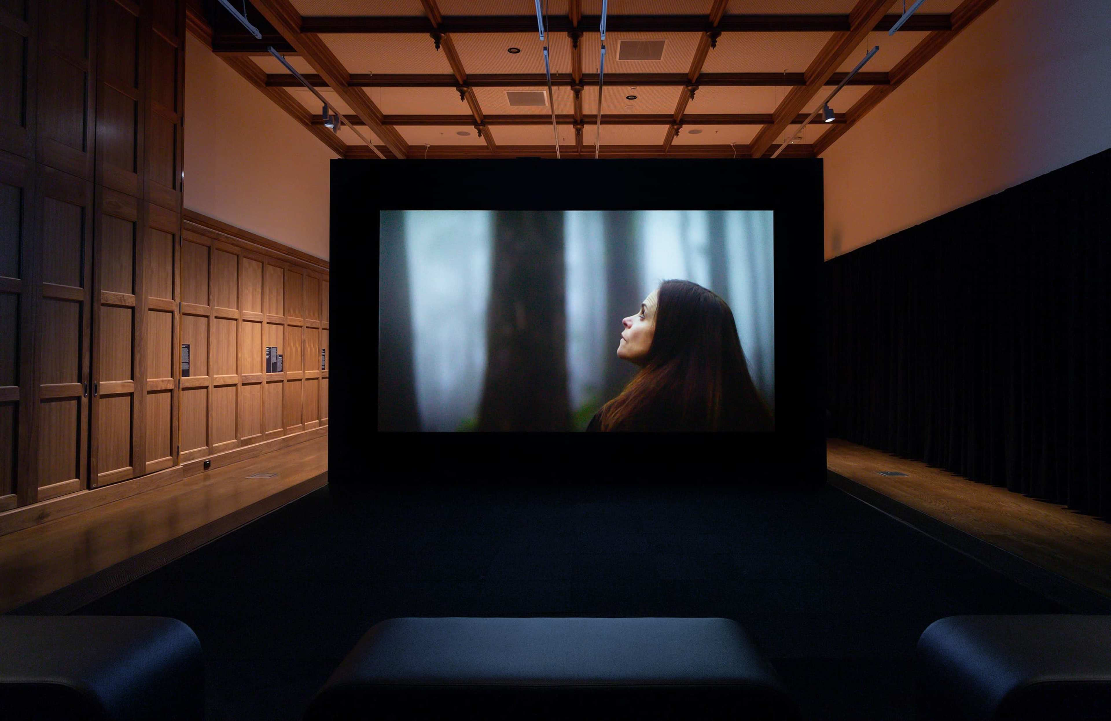 A dimly lit room with wooden paneling and black curtains features a large screen displaying a woman with long dark hair gazing upward in a misty forest. Benches are positioned in front of the screen for viewers.