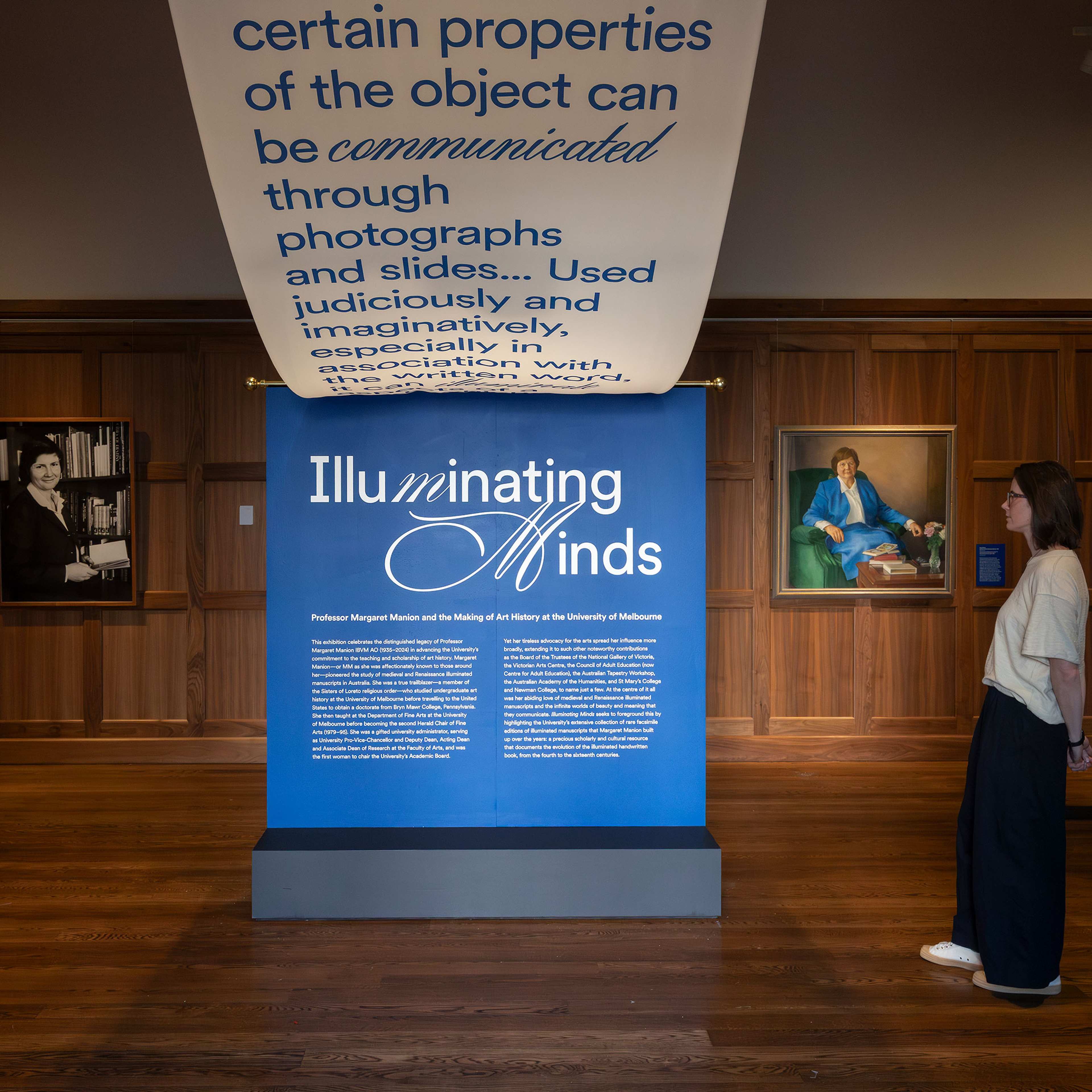 A person stands in a gallery, reading a blue display titled Illuminating Minds, with portraits on wood-panelled walls and a large banner with text hanging above.
