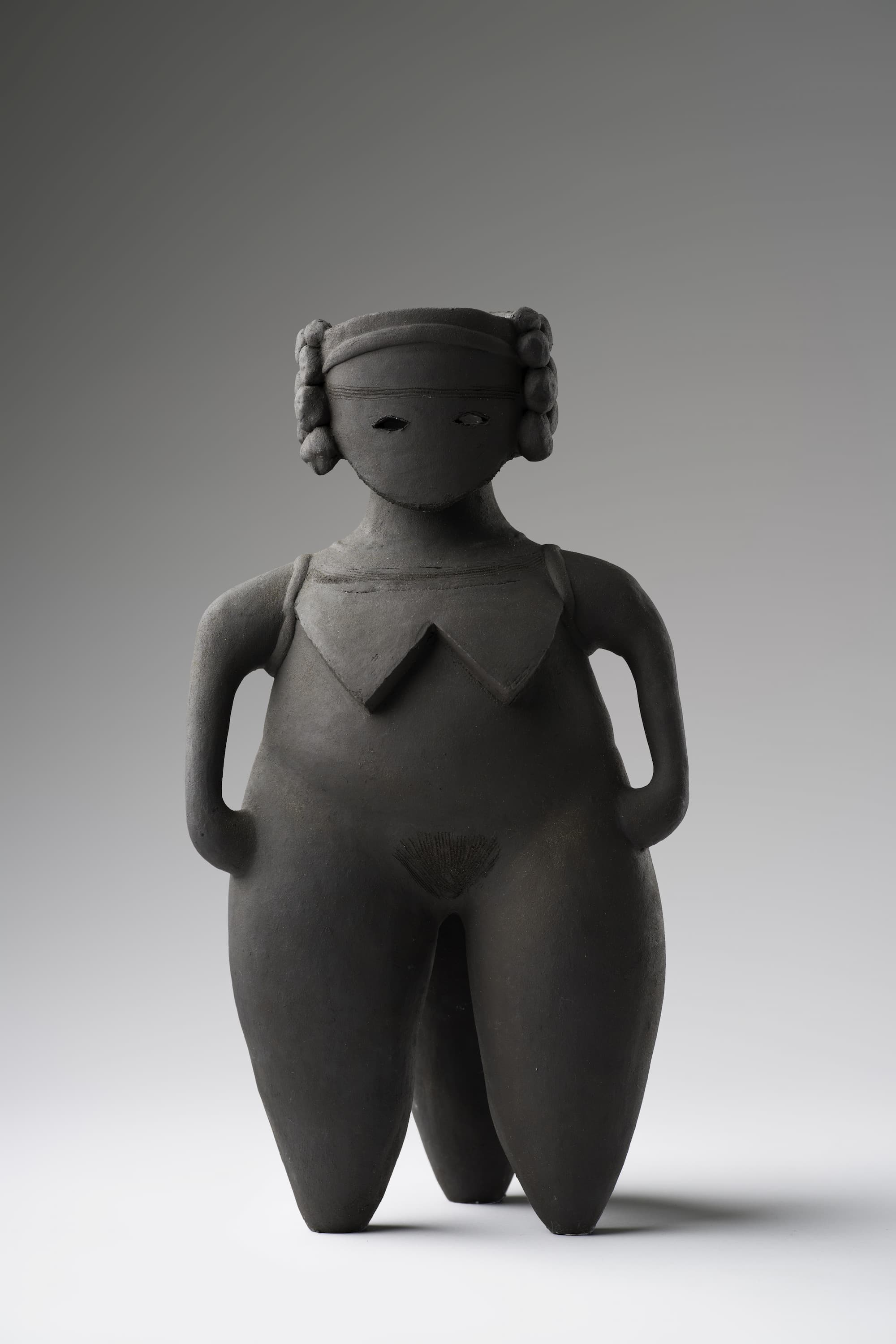A dark, stylized sculpture of a human figure with curly hair, minimal facial features, and broad limbs. The figure stands upright against a neutral background.