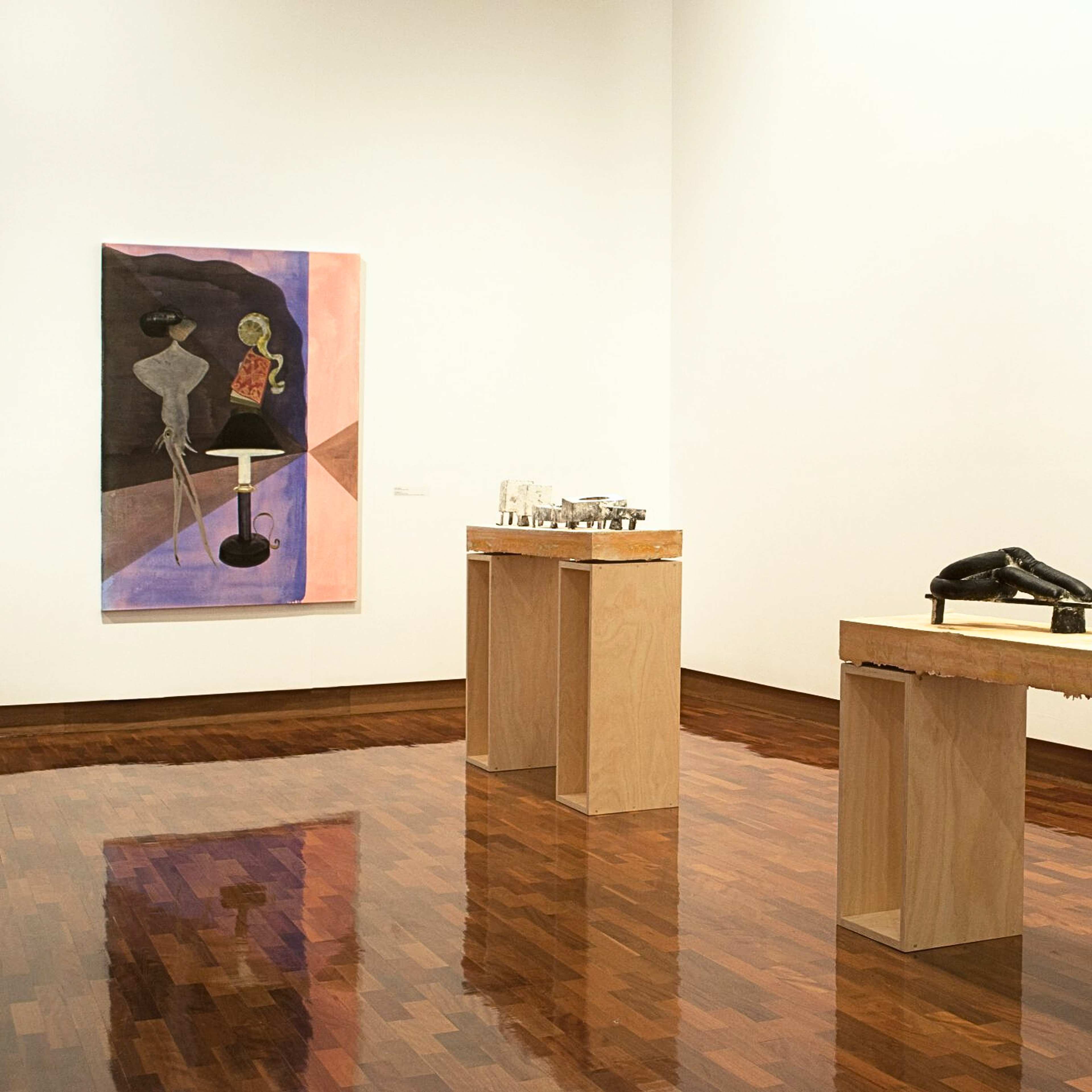 A photograph of a gallery with a painting on the left and sculptural works on large plywood plinths