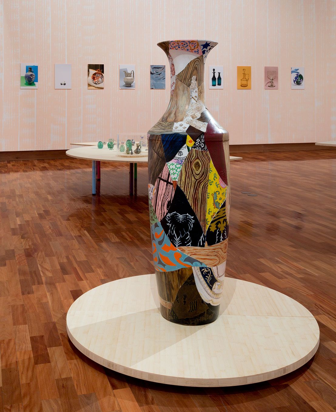 Largescale decorative colourful vase is displayed on plywood support with colourful prints hanging on the wall behind. 