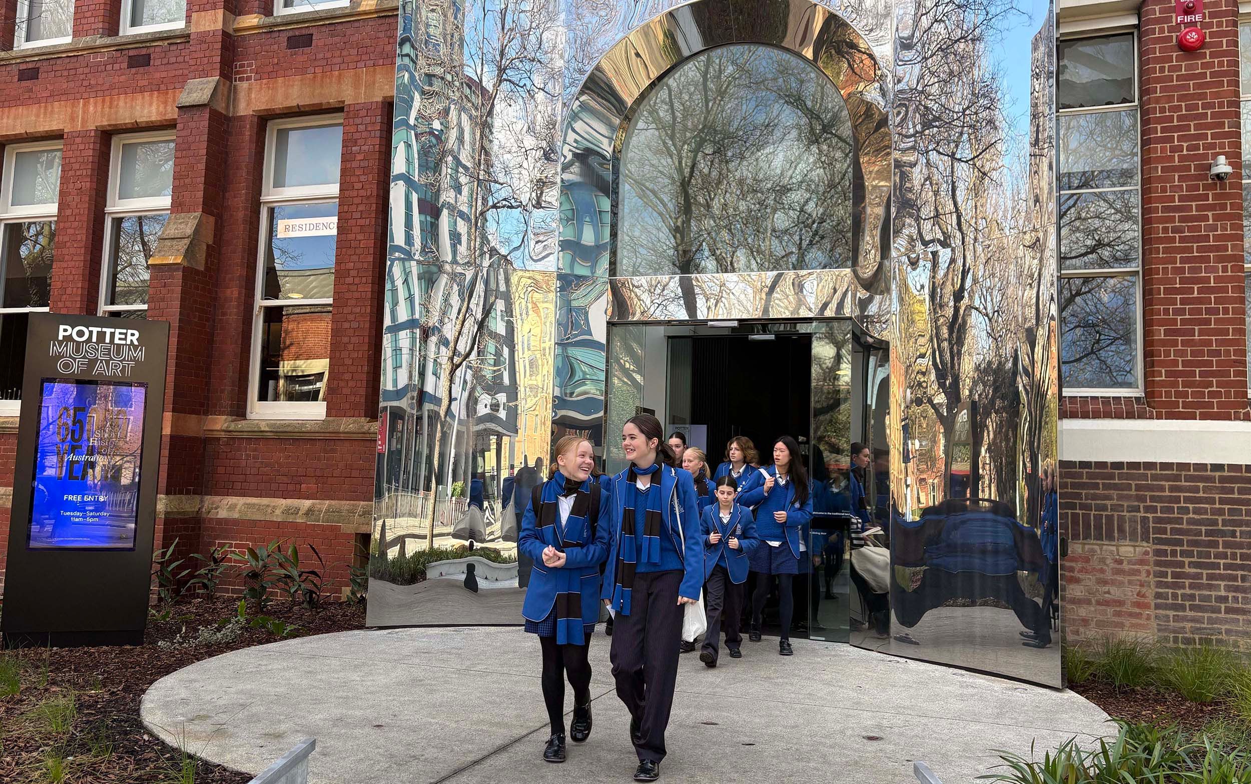A group of students in blue uniforms walk out of the glass entrance of the Potter Museum of Art, which reflects trees and nearby buildings. The museum’s exterior is a mix of brick and modern mirrored surfaces.