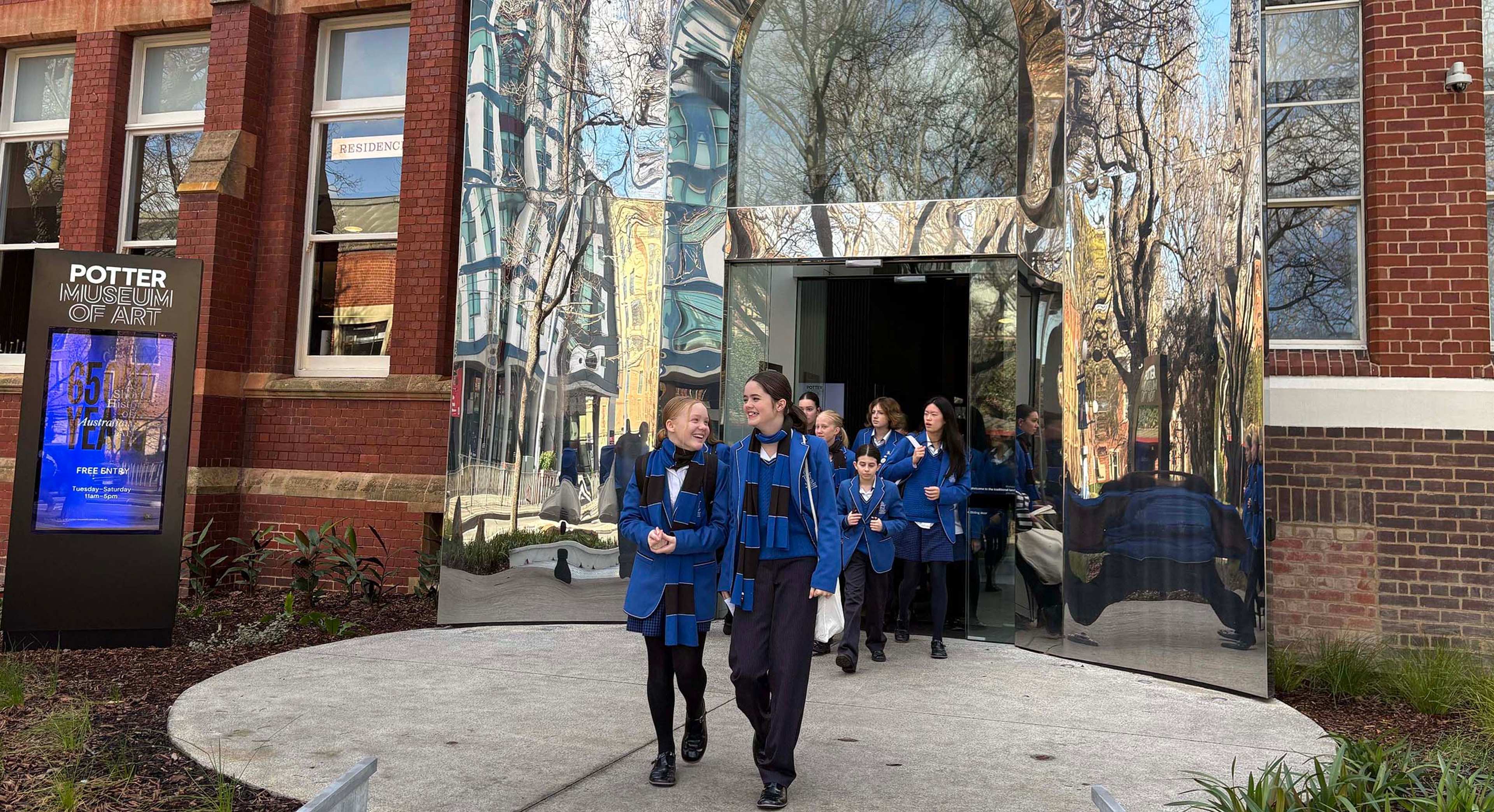 A group of students in blue uniforms walk out of the glass entrance of the Potter Museum of Art, which reflects trees and nearby buildings. The museum’s exterior is a mix of brick and modern mirrored surfaces.