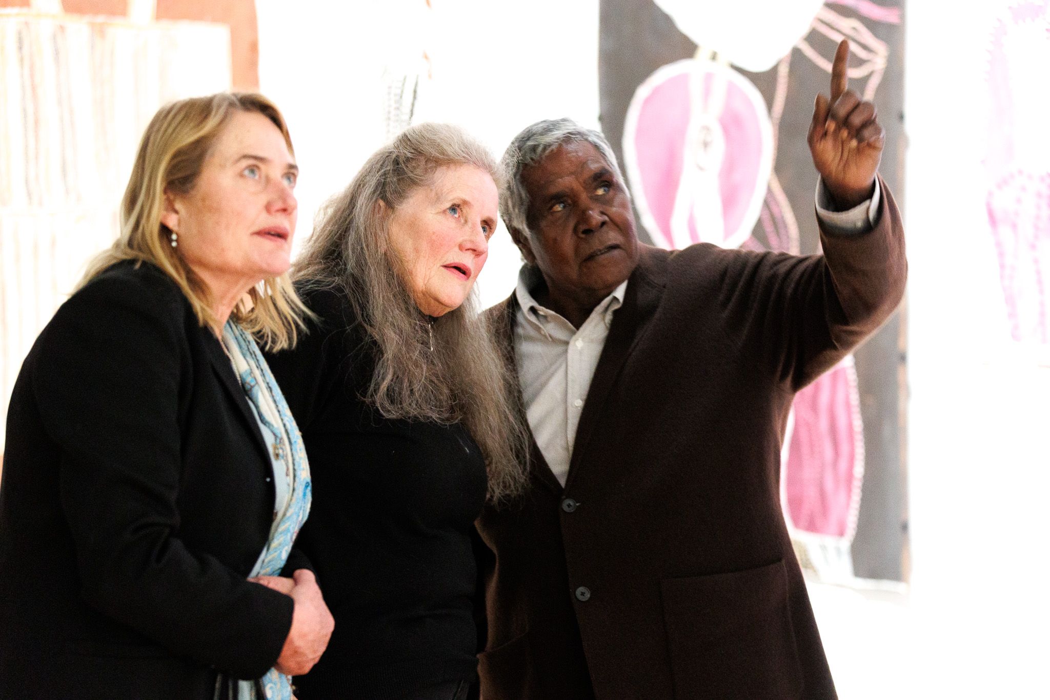 Three people stand closely together in an art gallery, looking intently in the same direction as a man on the right points upwards. Colourful abstract paintings are visible on the wall behind them.