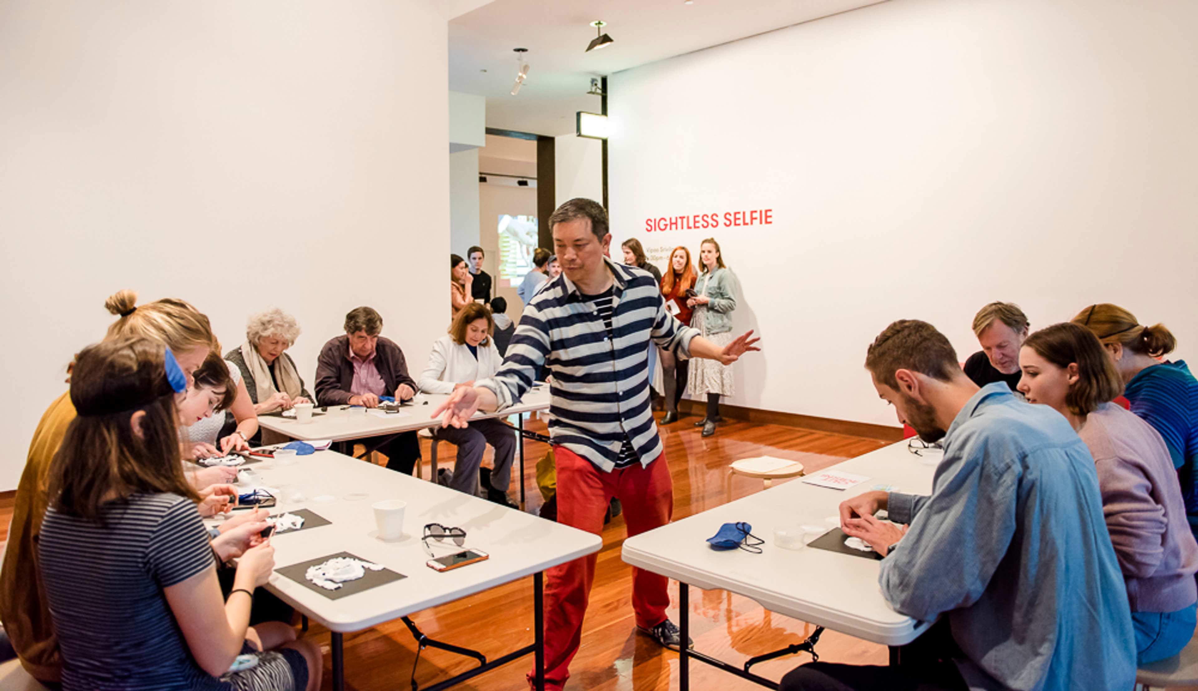 An artist engages with visitors at desk who make sculptures of themselves with blindfolds