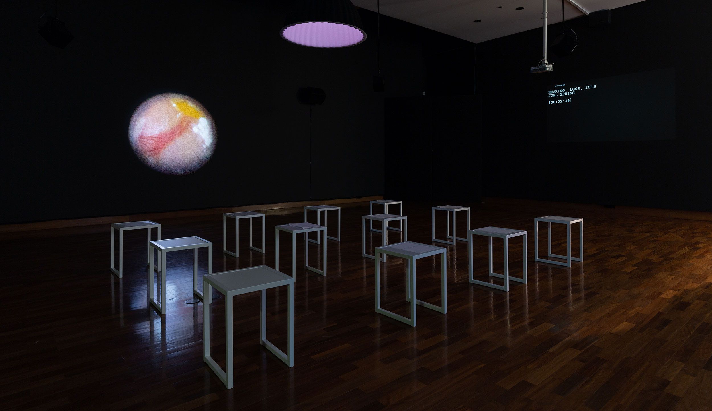 A darkened gallery space with white stools and two projected artworks, one displaying a text that  is hard to read and another an outline of a planet