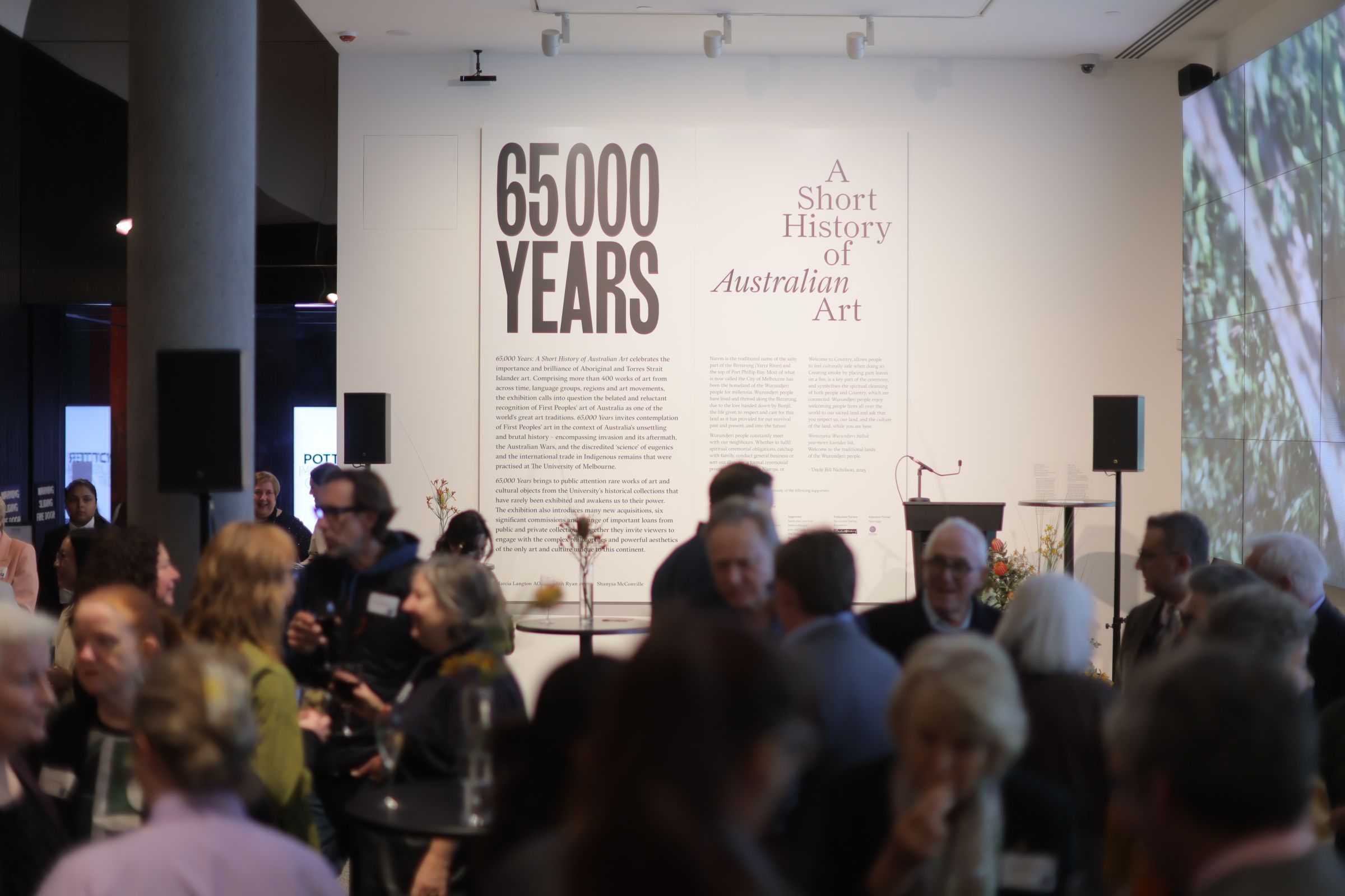 A group of people gather at an exhibition or event in front of a large wall display titled 65,000 Years: A Short History of Australian Art with text and flowers on a table beneath the sign.