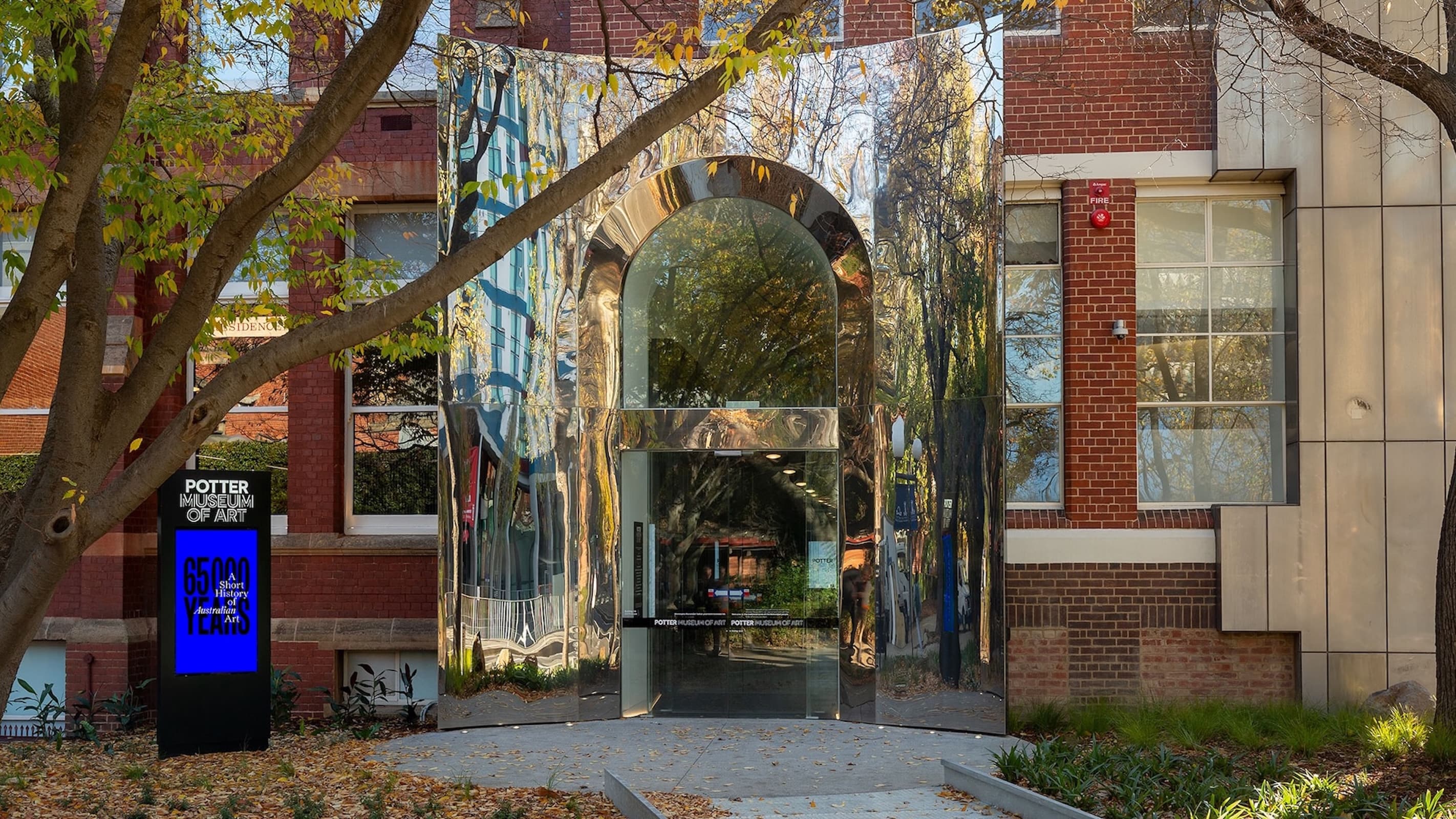 A modern mirrored glass entrance with an arched doorway contrasts with the red brick facade of the Potter Museum of Art. A digital sign stands on the left, surrounded by trees and autumn leaves on the ground.