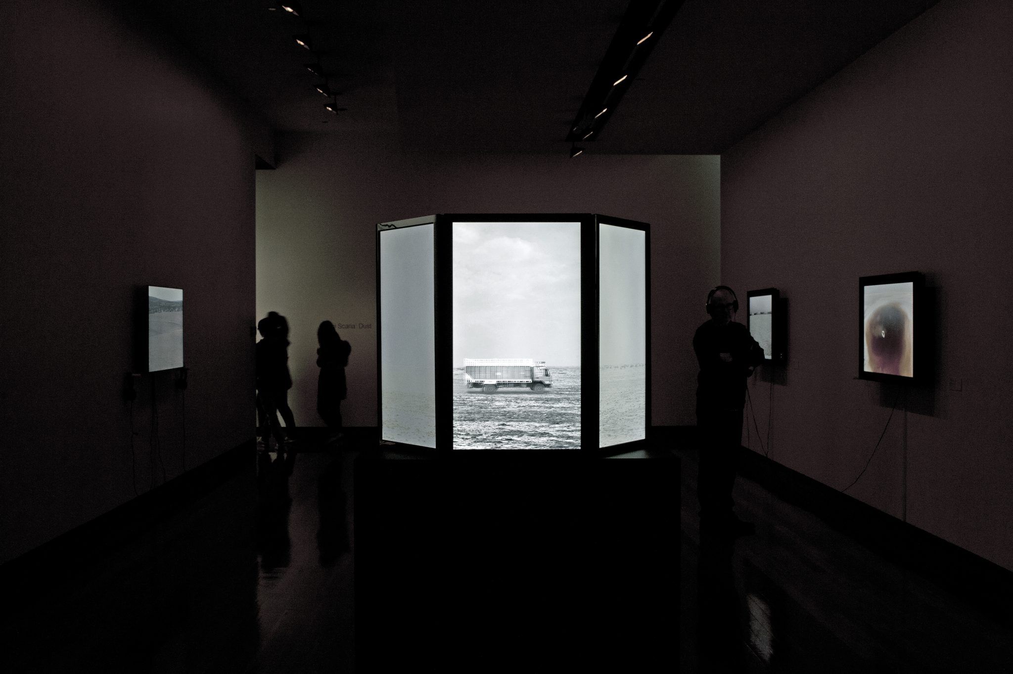 A photograph of a darkened gallery space with wall-mounted lightboxes 