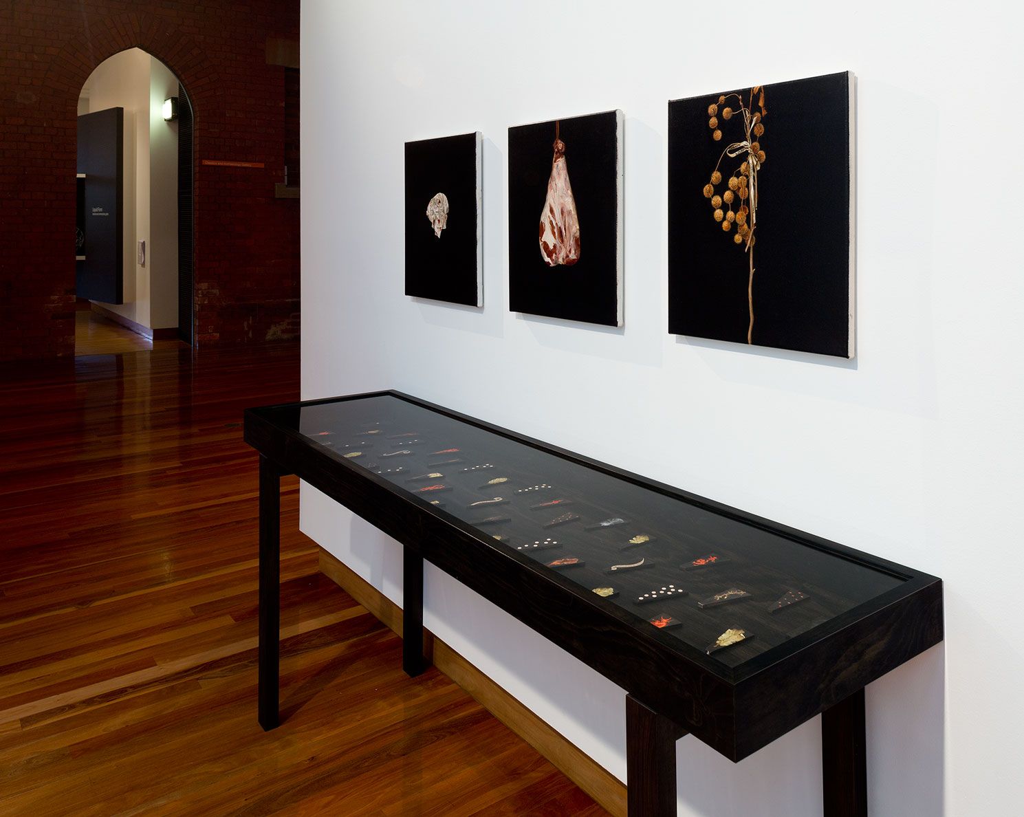 Three paintings in dark towns hang above a glass vitrine with small objects