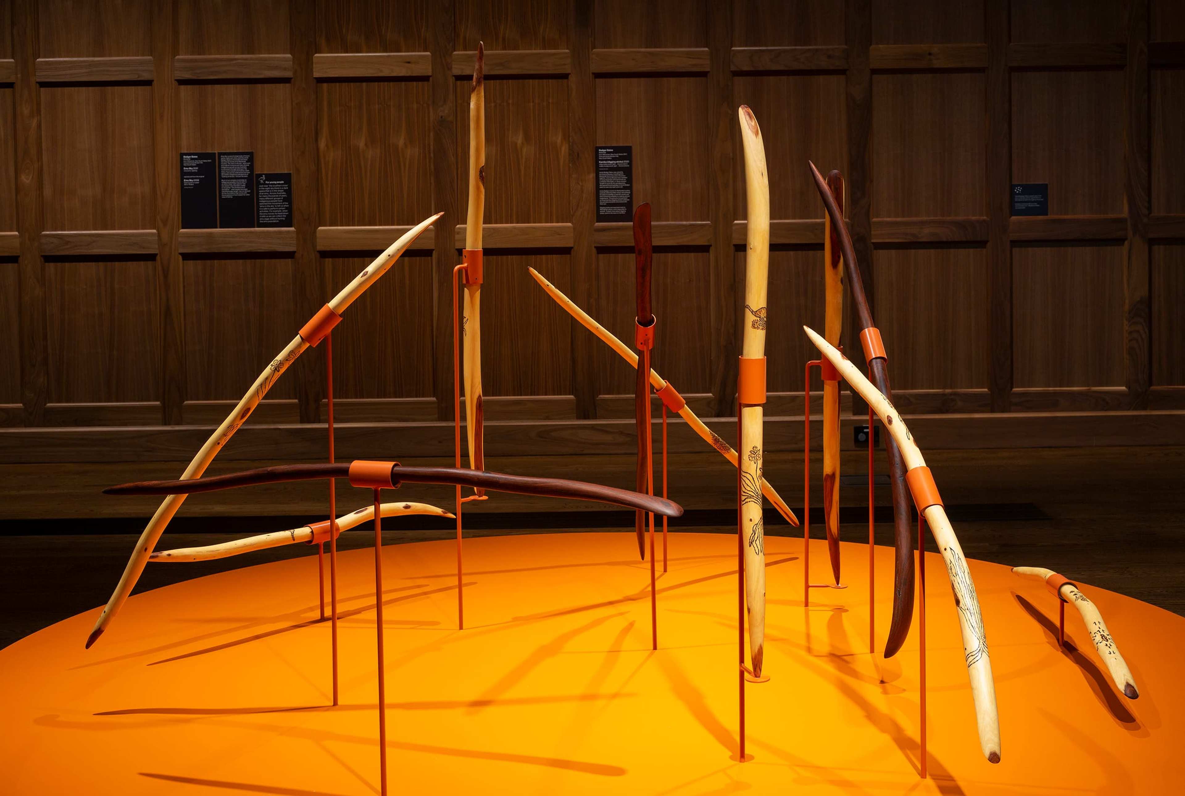 Curved wooden sticks of various sizes are displayed upright on an orange circular platform, each supported by orange stands, against a backdrop of dark wood paneling and exhibition placards.