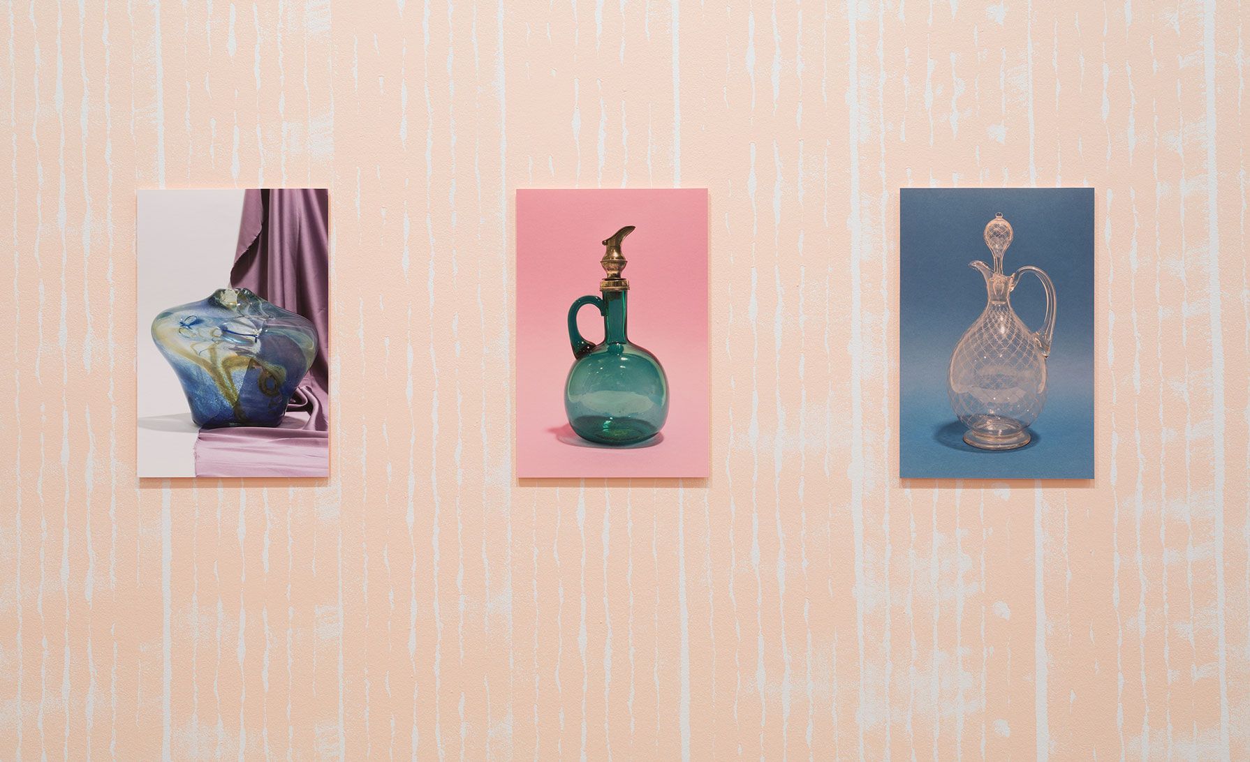 Three photographs of ornate glass jugs and a carafe hang in line on a pink wall
