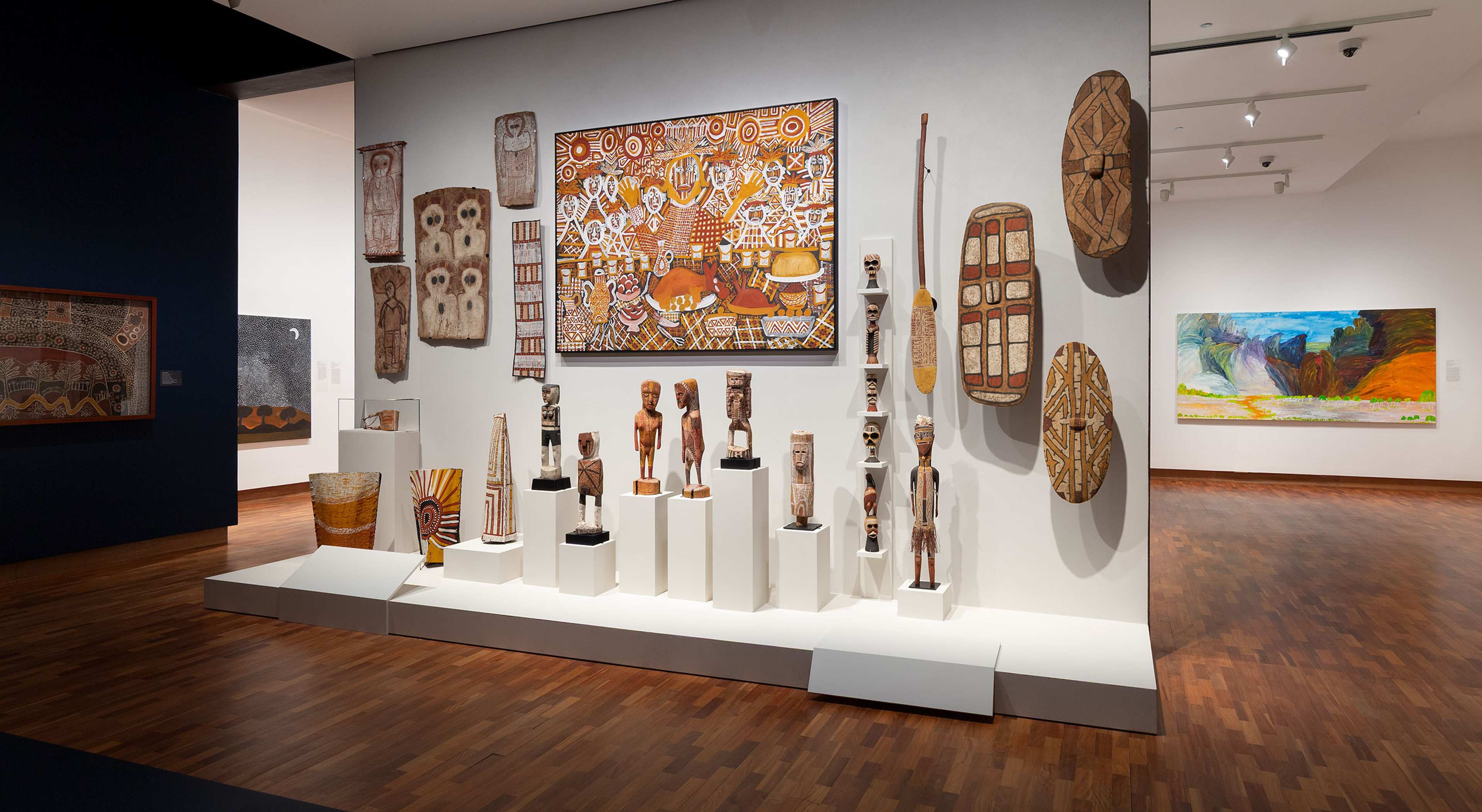 A museum gallery displays Indigenous Australian art, including painted wooden sculptures, shields, decorated panels, woven baskets, and a large, vibrant, abstract painting on the wall. The floor is polished wood.