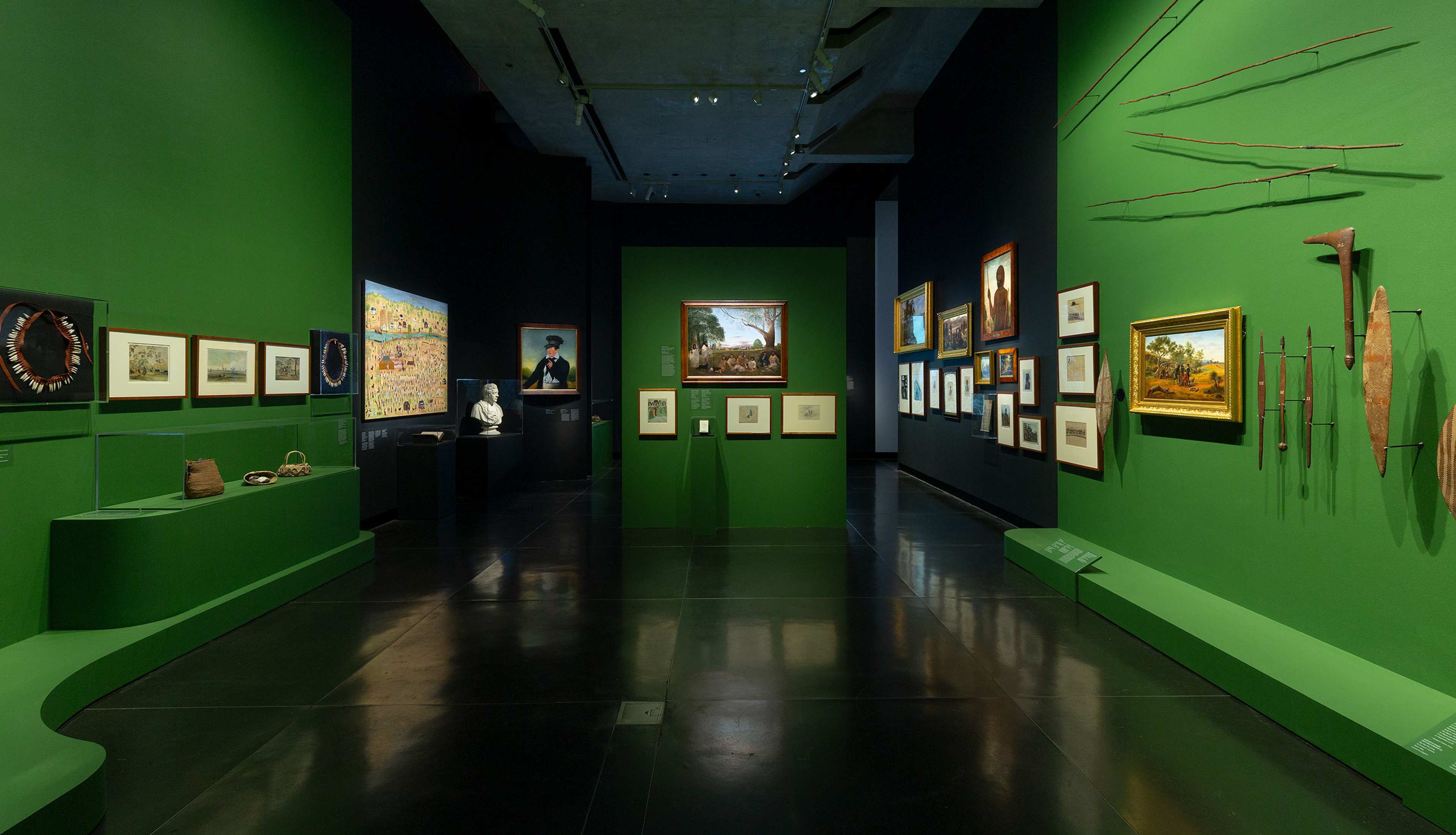 A museum gallery with green walls displays framed paintings, photographs, and sketches, as well as traditional tools and weapons. Sculptures and artifacts are also showcased on pedestals and shelves. The floor is glossy black.