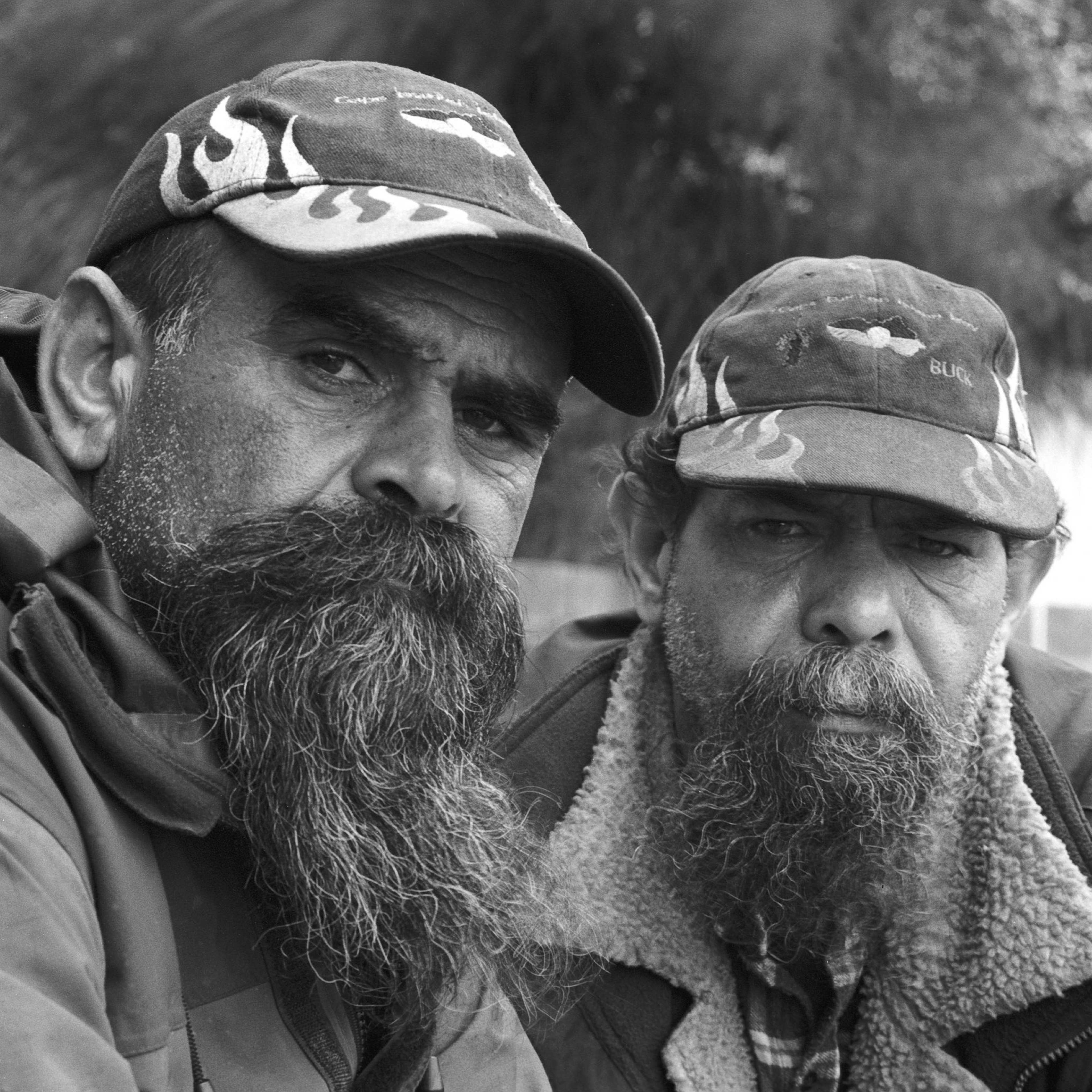 A black and white photograph showing faces of two people with beards wearing caps looking directly into the lens of the camera