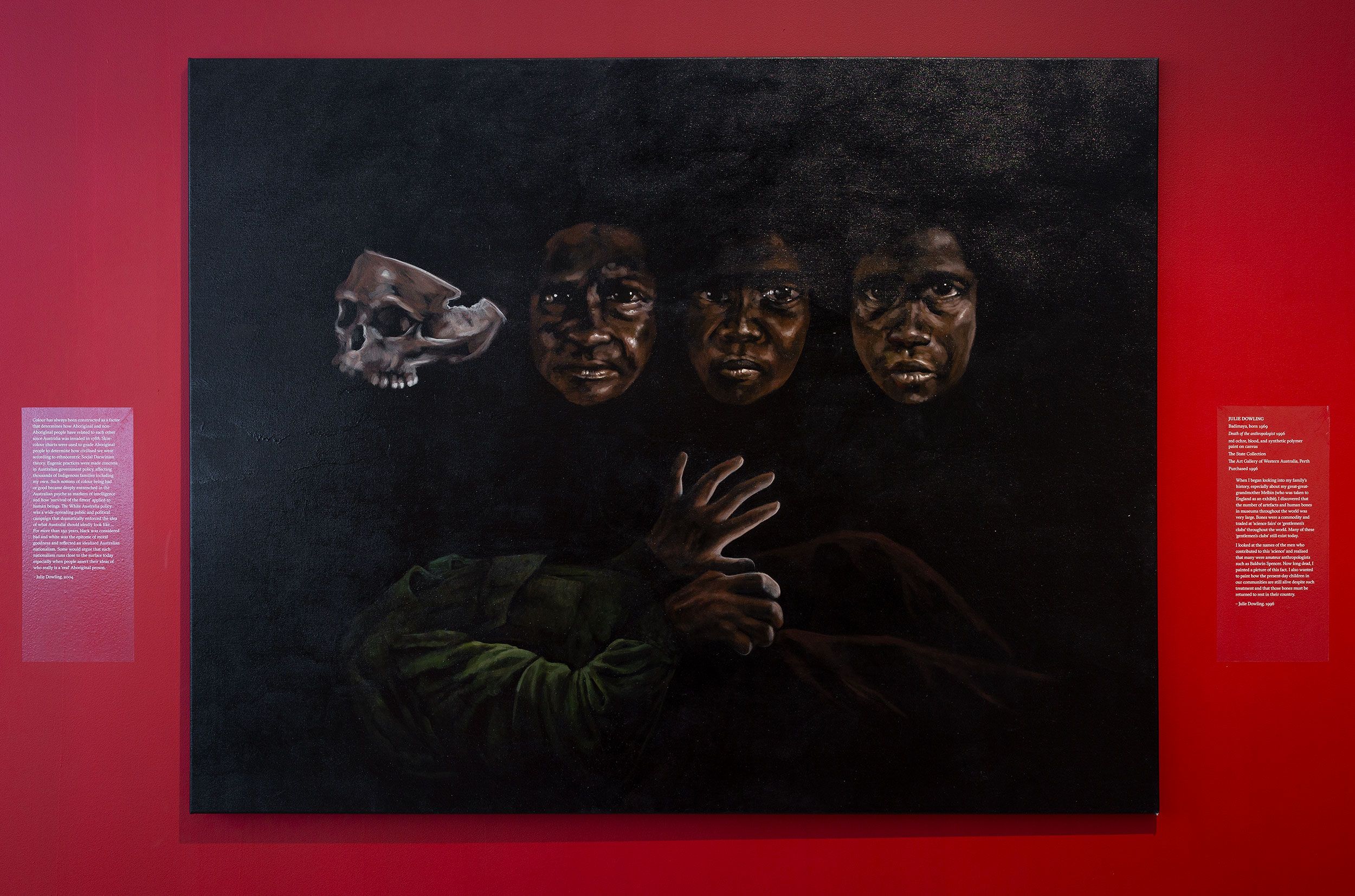 A painting on a red wall shows three dark-skinned faces and a hand emerging from shadows, with a skull to the left. The background is black, and two text plaques are displayed on either side of the artwork.