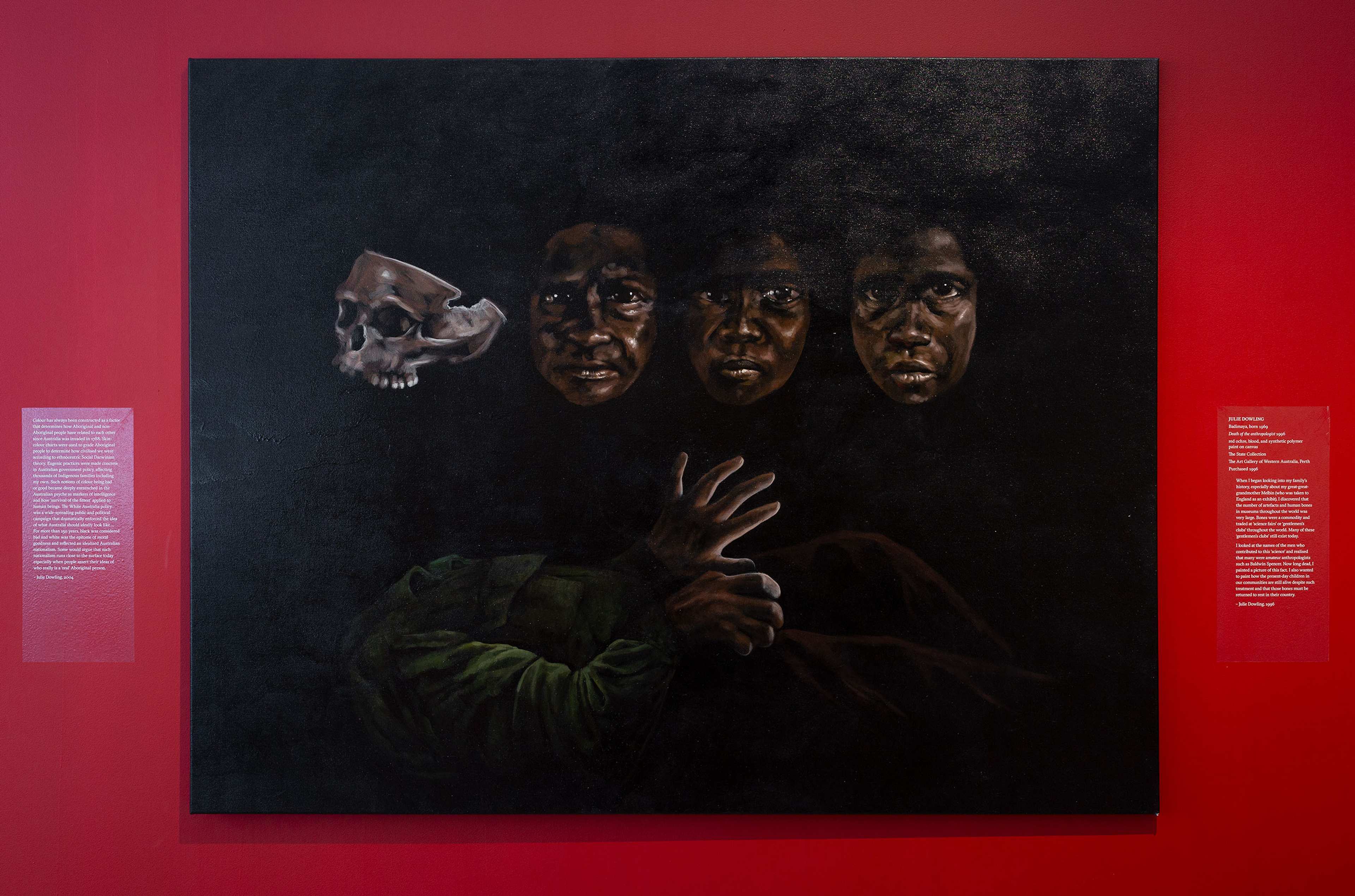 A painting on a red wall shows three dark-skinned faces and a hand emerging from shadows, with a skull to the left. The background is black, and two text plaques are displayed on either side of the artwork.