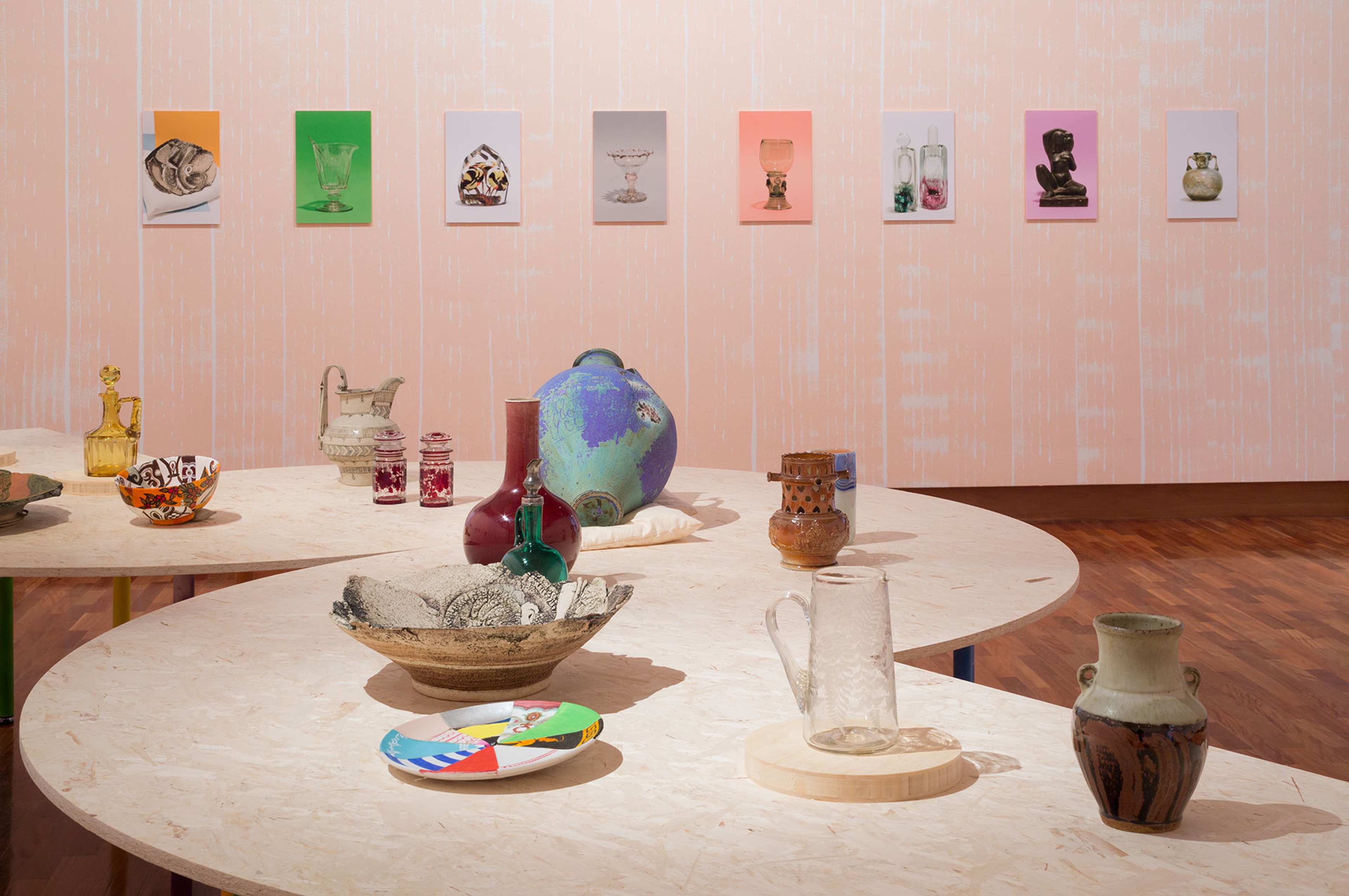 Plates bowls and decorative objects made from various materials are displayed on a plywood table with colourful prints hanging on the wall behind.
