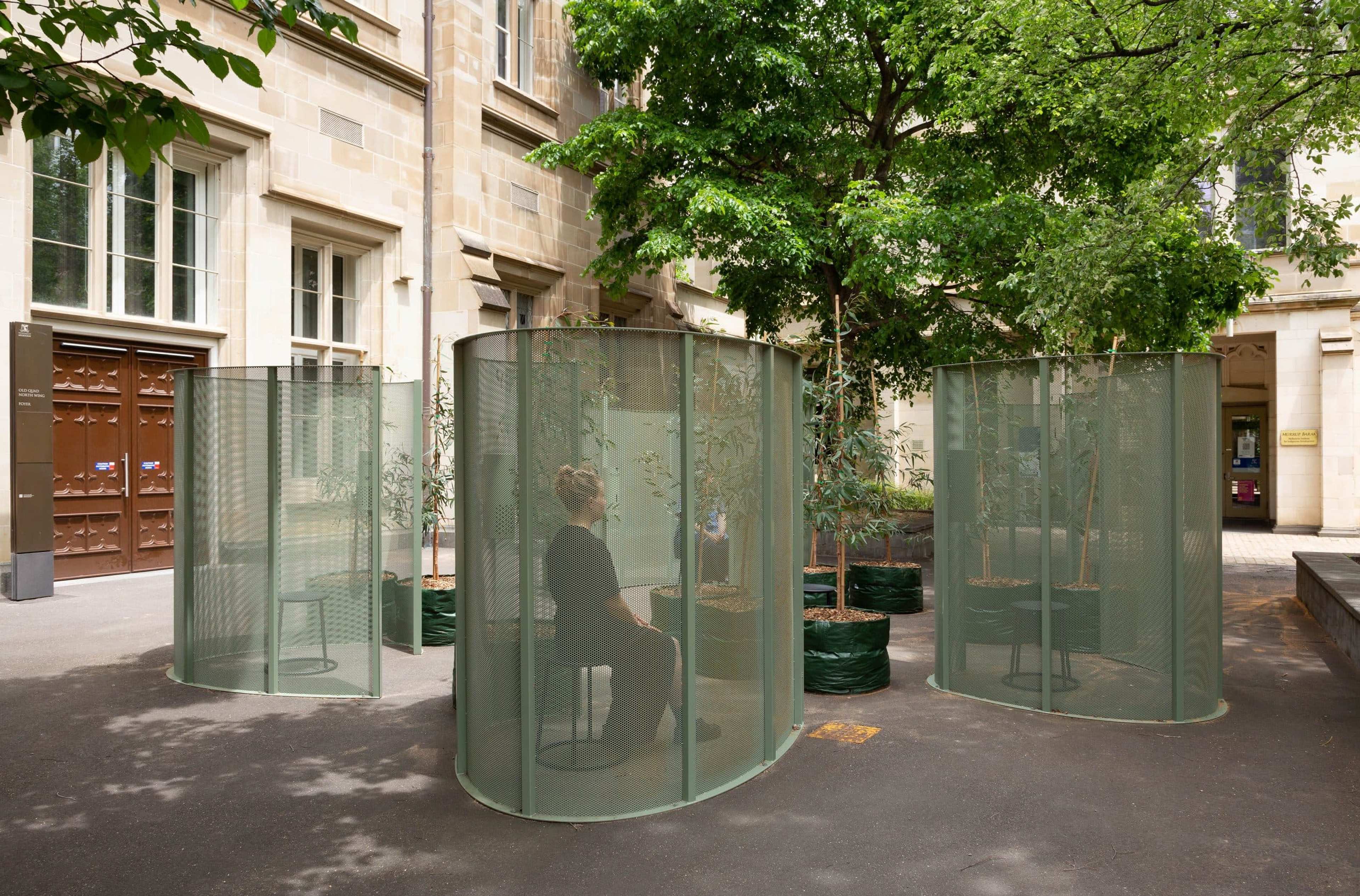 A person sits inside a circular green mesh enclosure among several similar structures in a tree-lined courtyard surrounded by stone buildings.