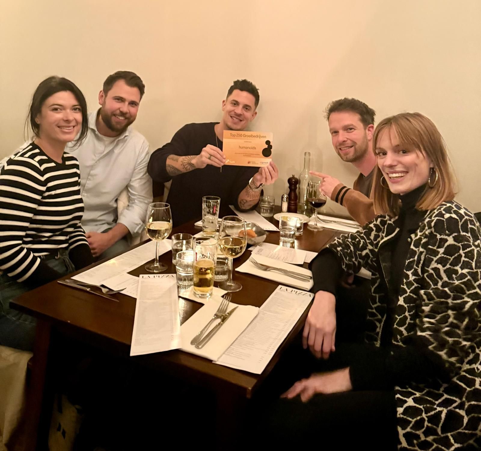 Het Humanoids team (Maxine, Wesley, Maarten, Robbin en Merel) zitten aan tafel in een restaurant. Maarten houdt een papier omhoog met daarop dat ze onderdeel zijn van de top 250 snelst groeiende bedrijven van Nederland.