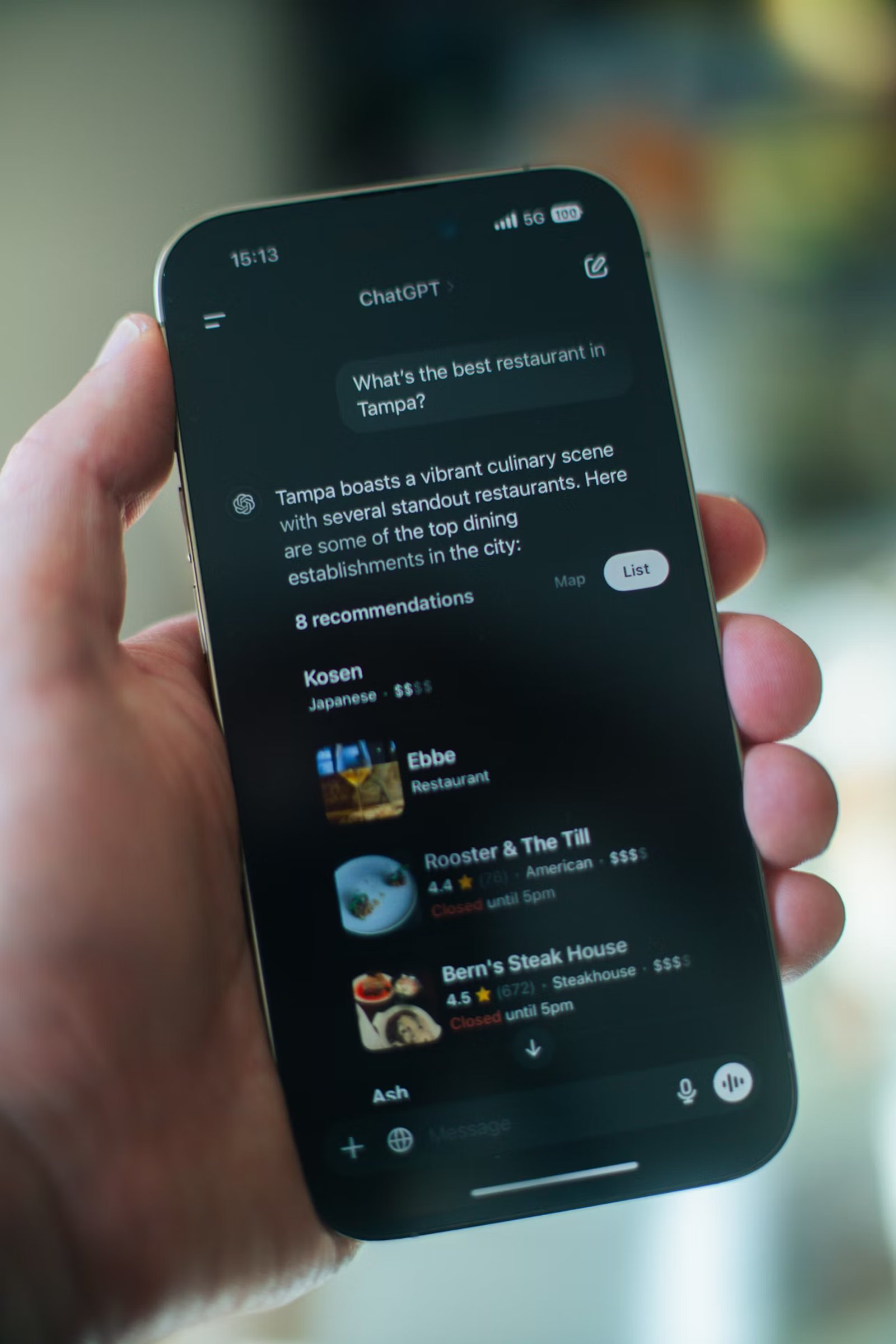 Een hand houdt een smartphone vast waarop de ChatGPT-app te zien is. Op het scherm staat de vraag “What’s the best restaurant in Tampa?” en de AI toont een lijst met restaurantaanbevelingen, inclusief namen, beoordelingen en openingstijden.