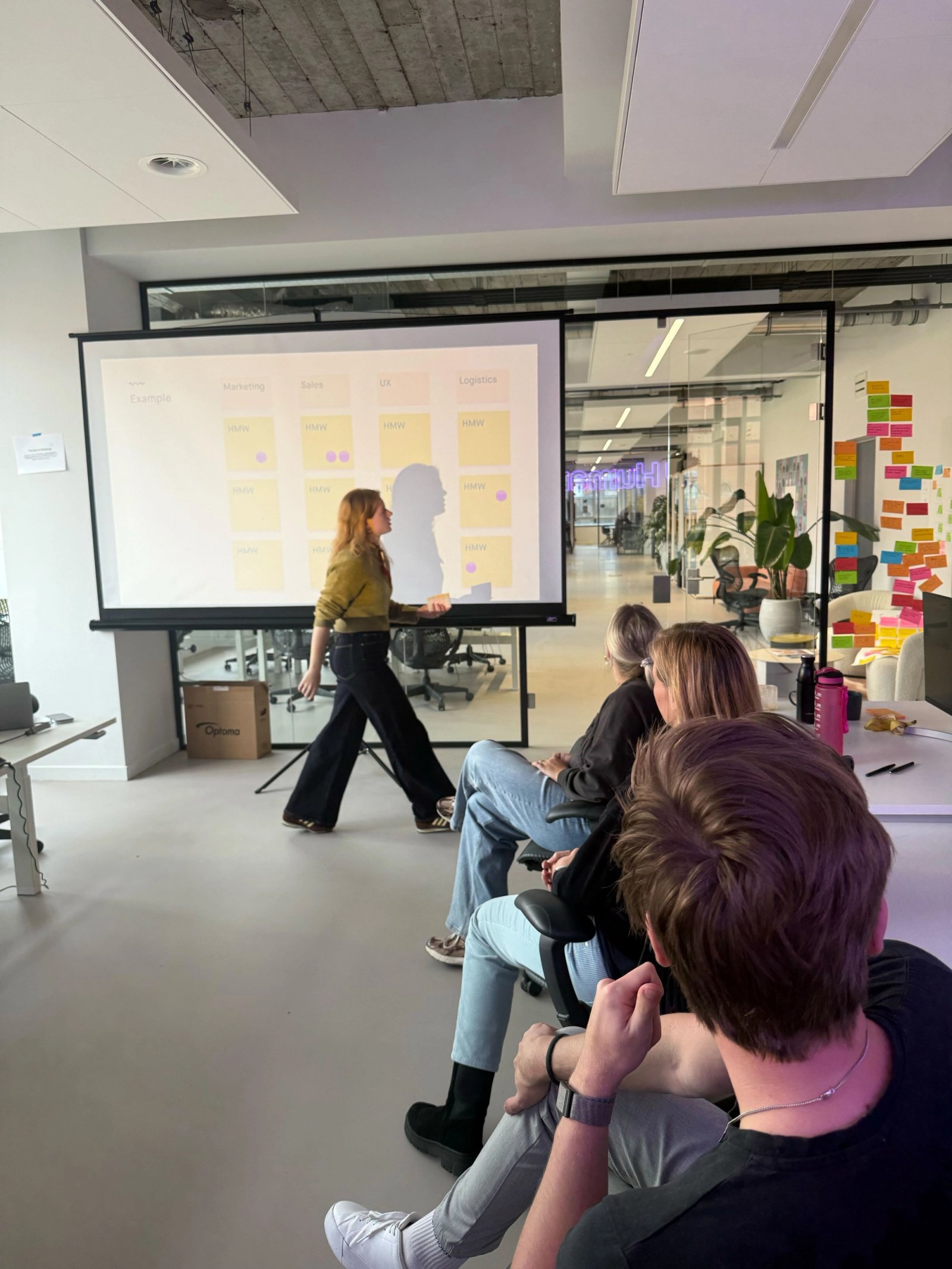 Een vrouw geeft een presentatie in een moderne kantoorruimte. Ze staat voor een groot scherm met post-its en slides, terwijl een kleine groep collega’s op stoelen toekijkt. Op de achtergrond zijn glazen wanden met meer post-its zichtbaar.