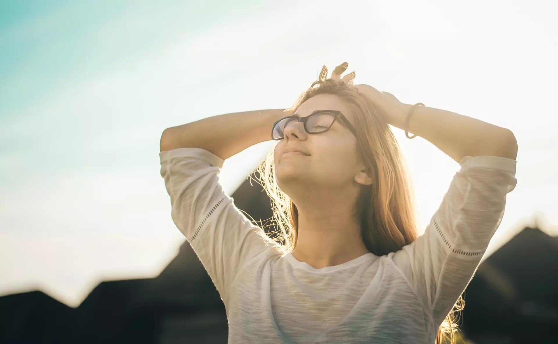 Una mujer con lentes relajada, manos en la cabeza y el sol a sus espaldas.