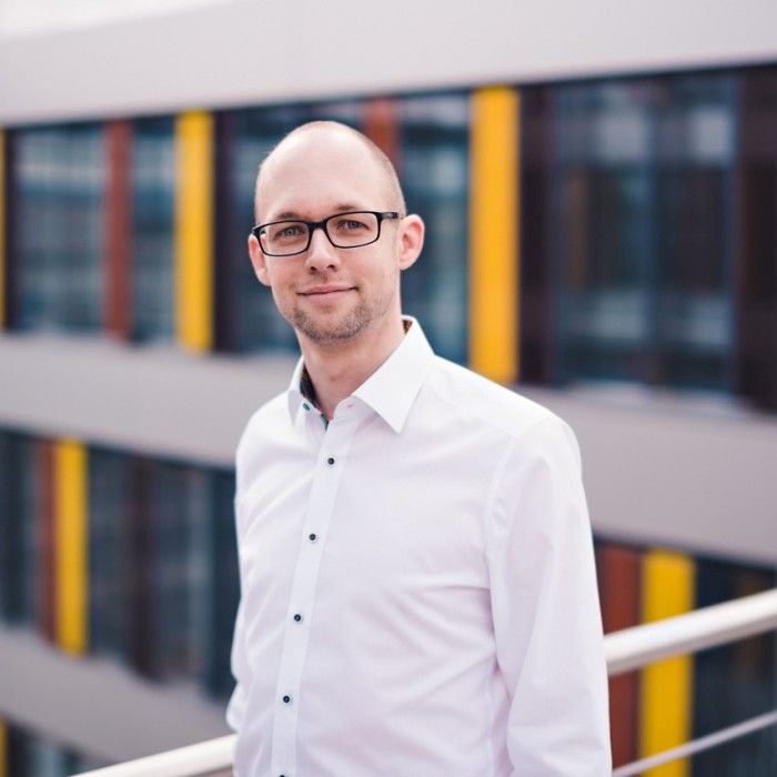 Blonder Mann mit Brille und weißem Hemd steht vor einem Bürogebäude mit gelben und roten Akzenten.