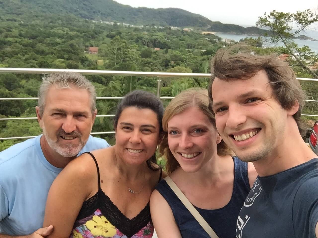 Vier lächelnde Personen nehmen ein Selfie vor einer grünen Landschaft mit Bergen und Wasser im Hintergrund.