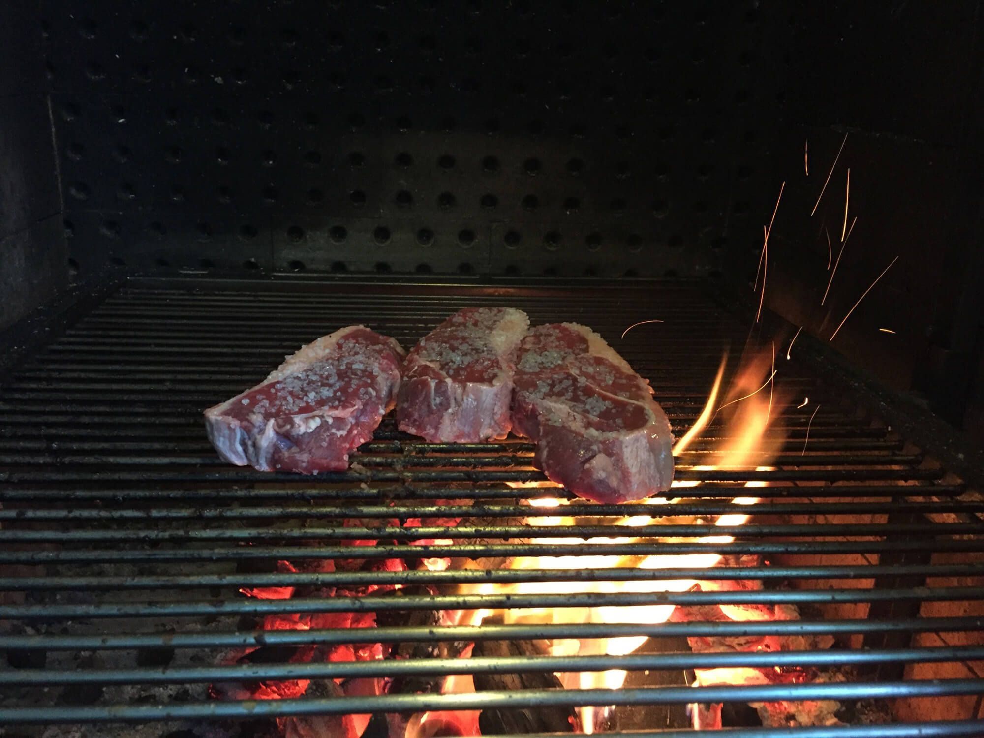 Drei mit Salz bestreute Steaks liegen auf einem Grillrost über glühender Holzkohle und Flammen.